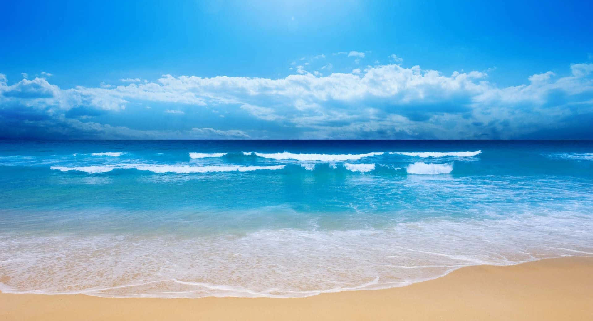 多雲天空下的可愛夏日海灘 壁紙 — 陽光在碧綠色的海浪上閃爍，海浪拍打著金色的沙灘，上方是廣闊的藍天和蓬鬆的白雲。