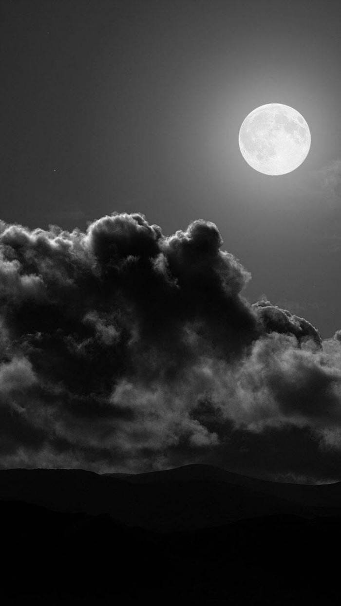 多雲純黑 iPhone 壁紙 — 一輪明亮的滿月在單色調的山脈背景下，在戲劇性的風暴雲剪影上方發光。