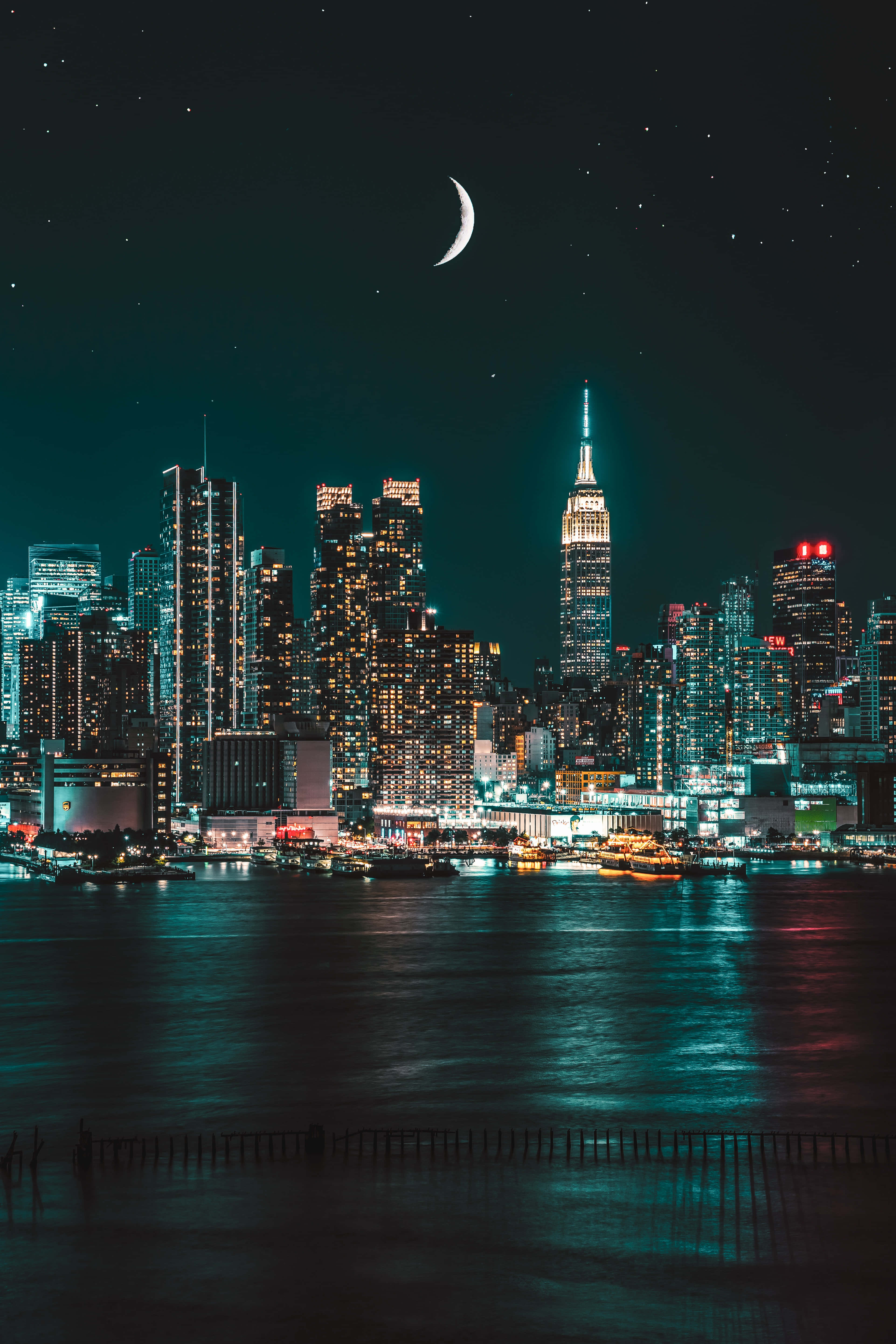 夜城霓虹閃耀的街道 壁紙 — 一彎新月懸掛在閃爍的夜景城市上空，摩天大樓倒映在暗水中。