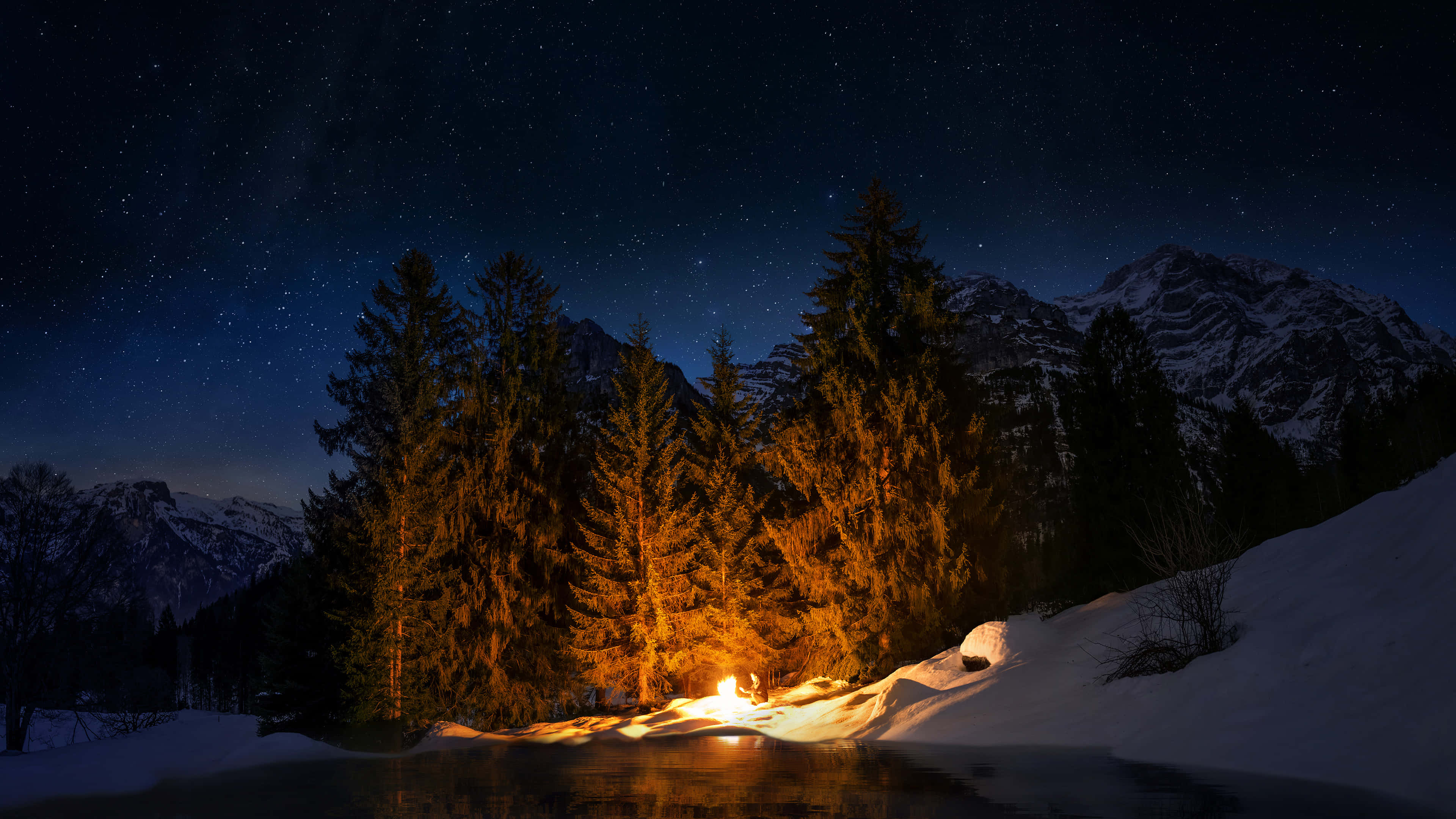 夜山營火 壁紙 — 在星空下，一個孤獨的營火在雪湖旁溫暖地燃燒，周圍有松樹和遠山。
