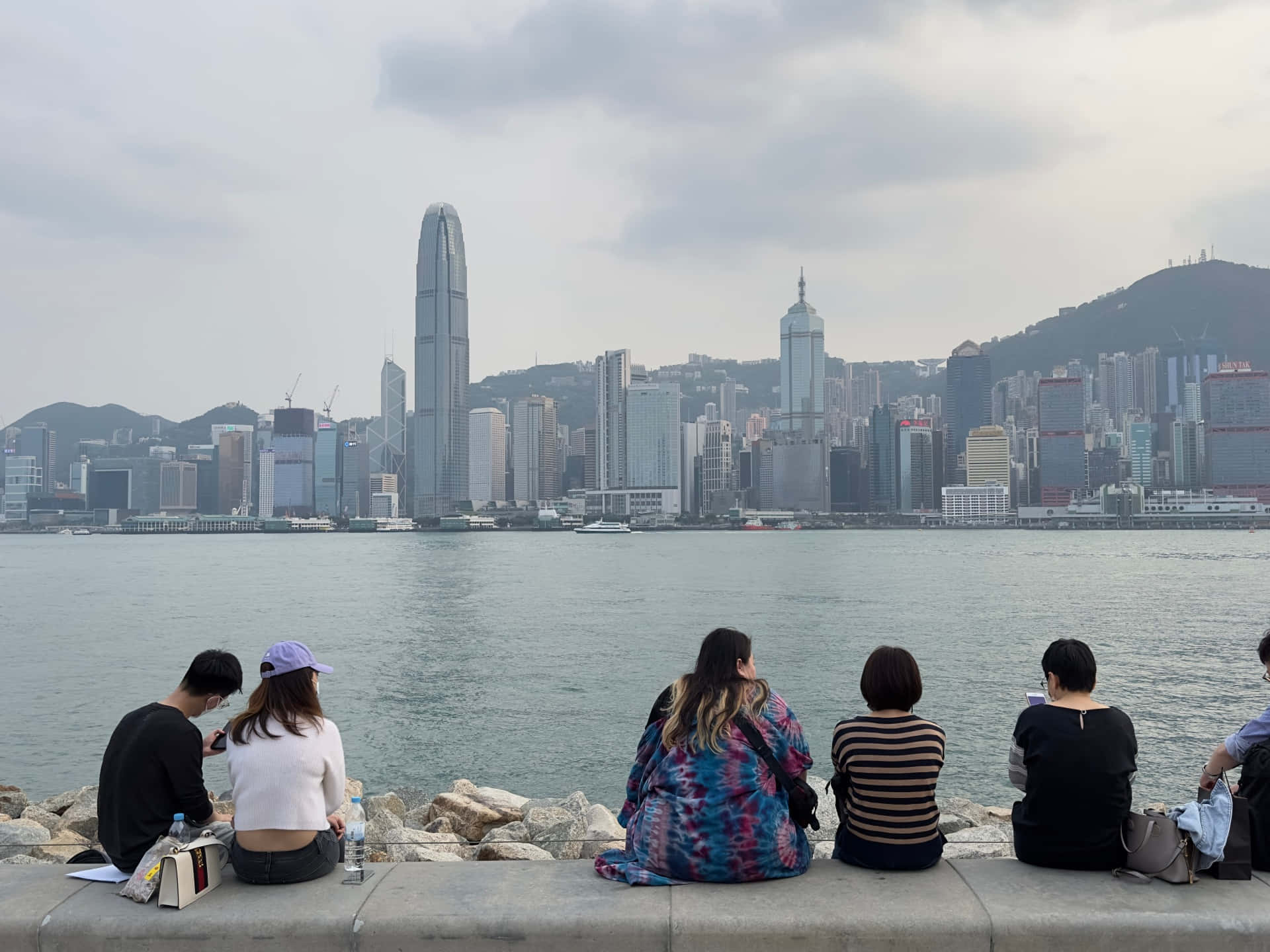 夜幕下的香港城市天際線 壁紙 — 五個人坐在石壆上，俯瞰陰天下香港標誌性的天際線和港口。