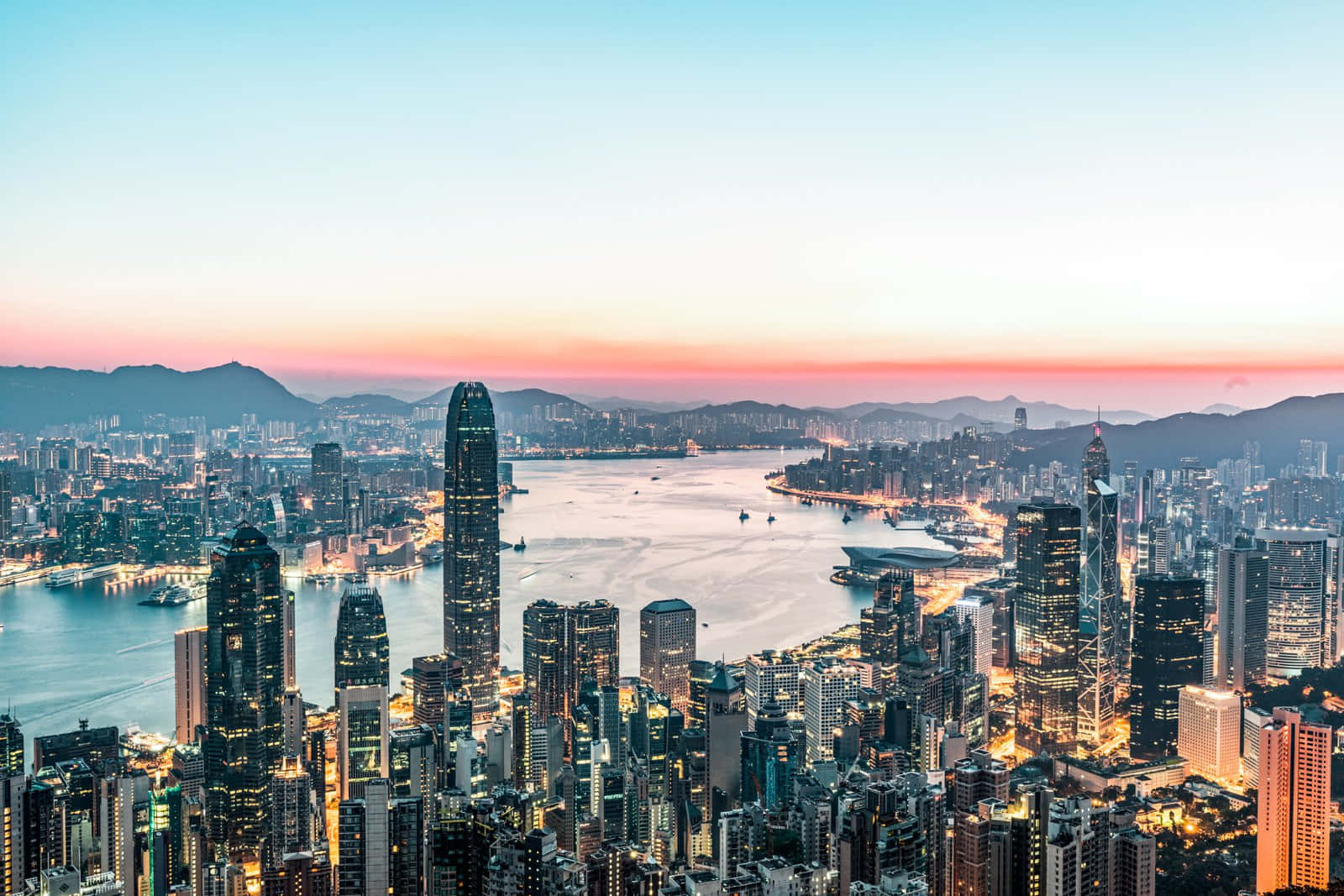 夜幕下的香港摩天大樓令人歎為觀止 壁紙 — 香港天際線的 panoramic 景觀，夕陽下閃爍的摩天大樓和寧靜的海港反射著粉紅色和藍色的天空。