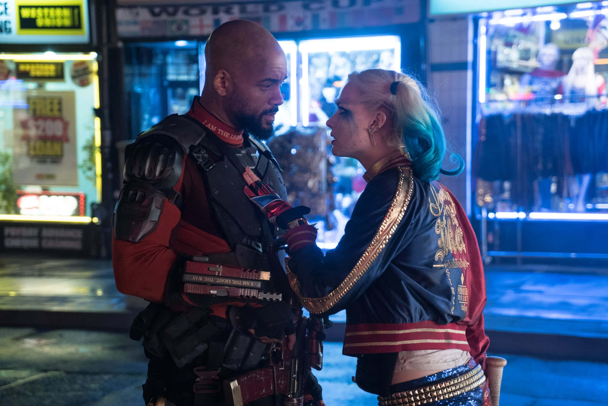 Deadshot And Harley Quinn 桌布 壁紙 — 夜晚，一個光頭男子身穿戰術裝備，與一位帶有藍色挑染的金髮女子在霓虹燈照亮的城市小巷中緊張對峙。
