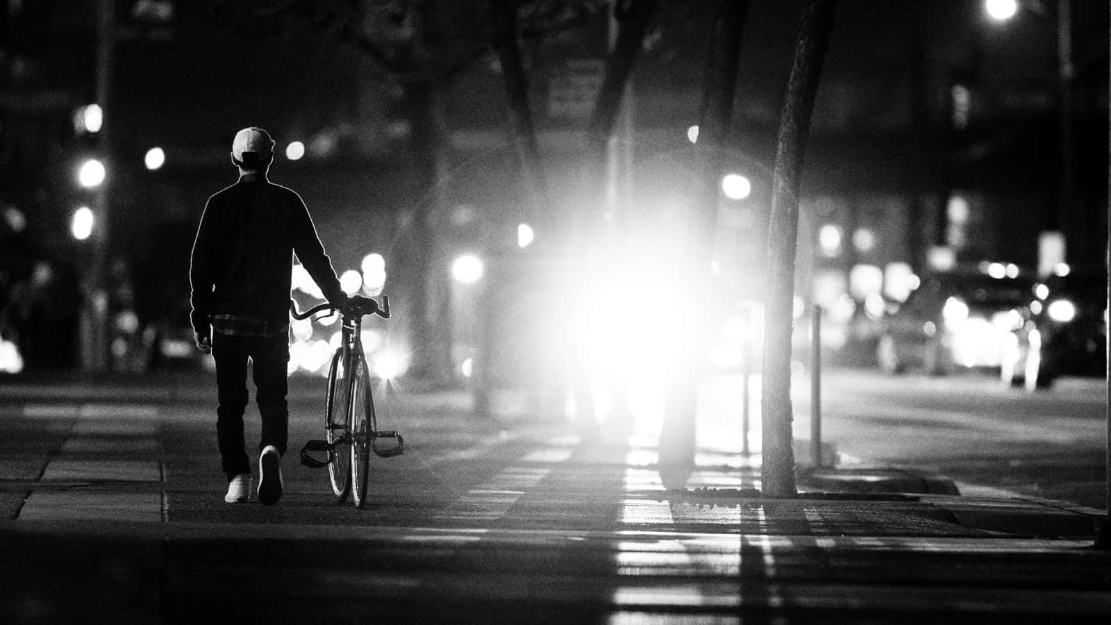 Black And White Street 桌布 壁紙 — 夜晚，一個孤獨的騎單車者推著單車走在城市街道上，明亮的車頭燈在前方形成發光的霧氣。