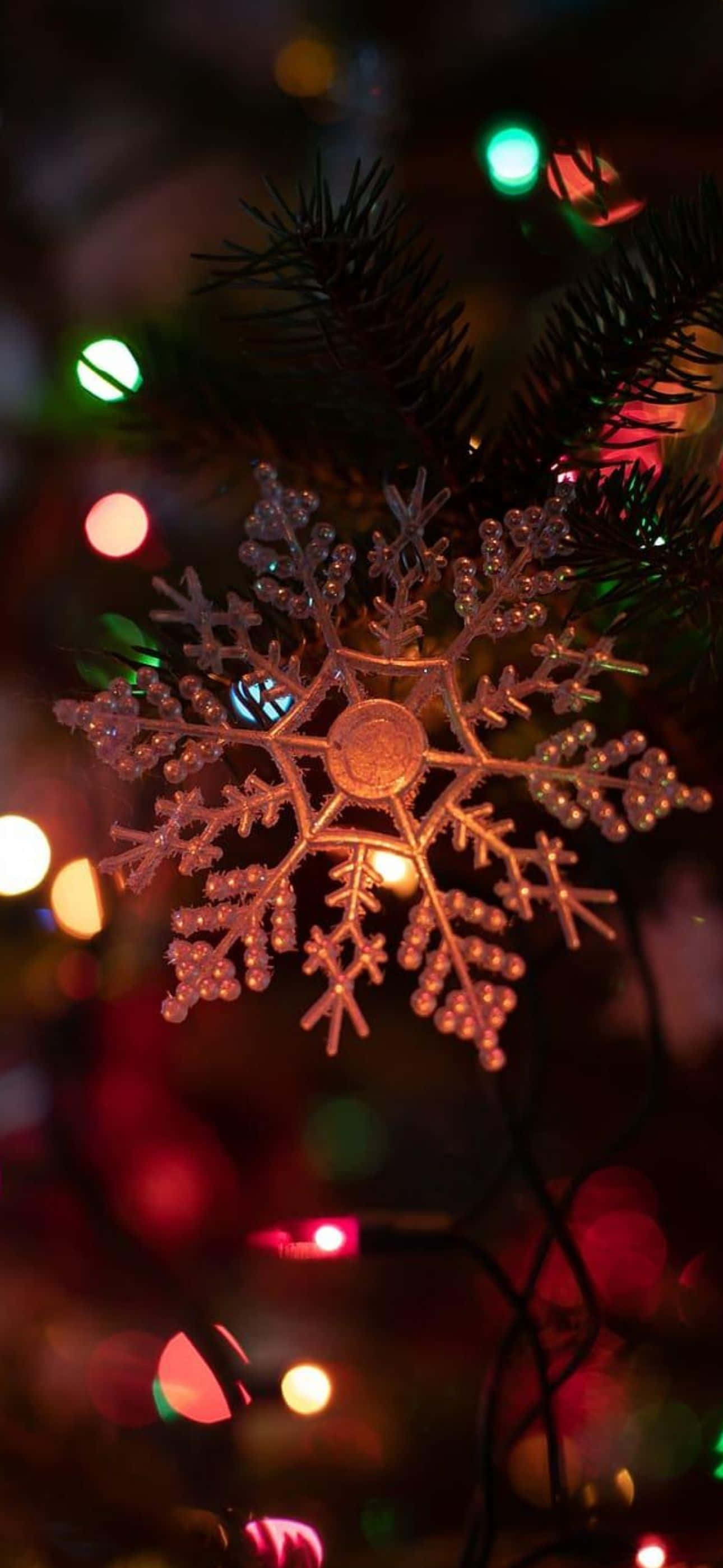 聖誕節 iPhone 桌布 壁紙 — 夜晚，一個閃閃發光的雪花裝飾品掛在松樹枝上，被色彩繽紛的散景燈光照亮。