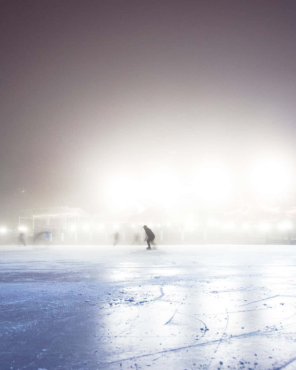 Ice Rink 桌布 壁紙 — 夜晚，一名孤獨的溜冰者在被迷霧籠罩的明亮體育場燈光下滑過冰凍的溜冰場。
