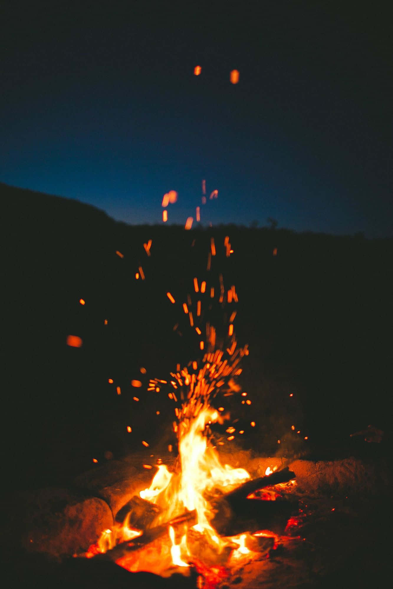 Beach Bonfire 桌布 壁紙 — 夜晚一個充滿活力的營火，讓發光的餘燼在深藍色的暮色天空中飛舞。