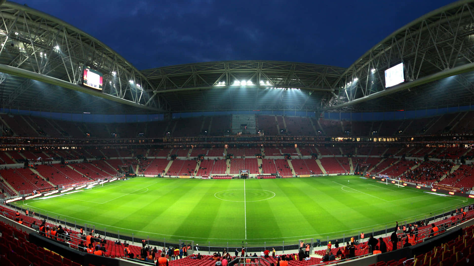 Soccer Stadium 桌布 壁紙 — 夜晚一個廣闊空蕩的足球場，明亮的泛光燈照亮了充滿活力的綠色球場和一行行紅色座位，上方是漆黑的天空。