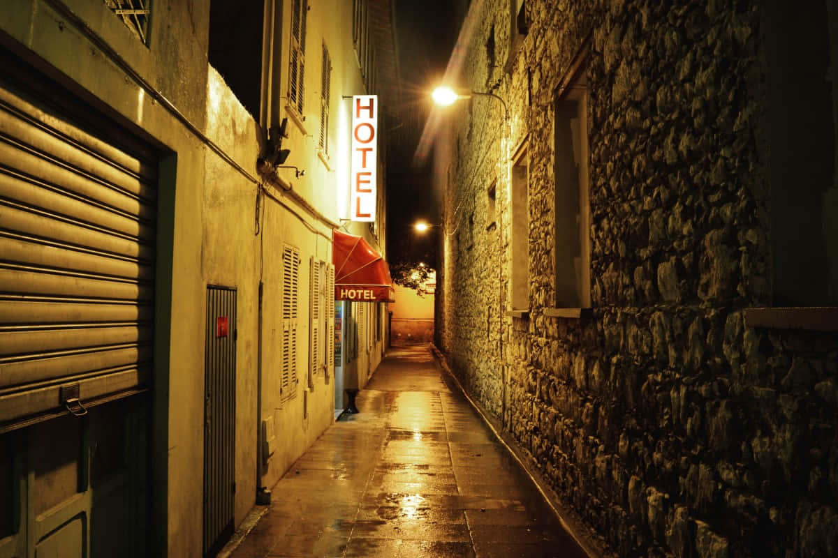 Alleyway 桌布 壁紙 — 夜晚一條狹窄潮濕的小巷，被發光的酒店招牌和路燈照亮，倒映在潮濕的鵝卵石上。