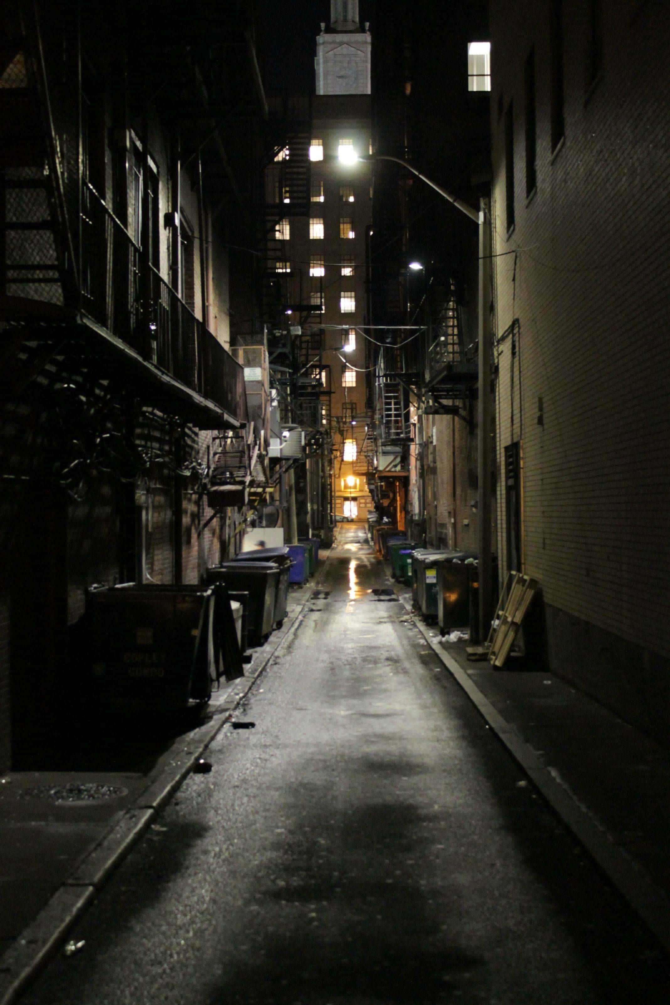Alleyway 桌布 壁紙 — 夜晚一條狹窄潮濕的小巷，被街燈和遠處建築物的窗戶照亮，投下長長的影子。