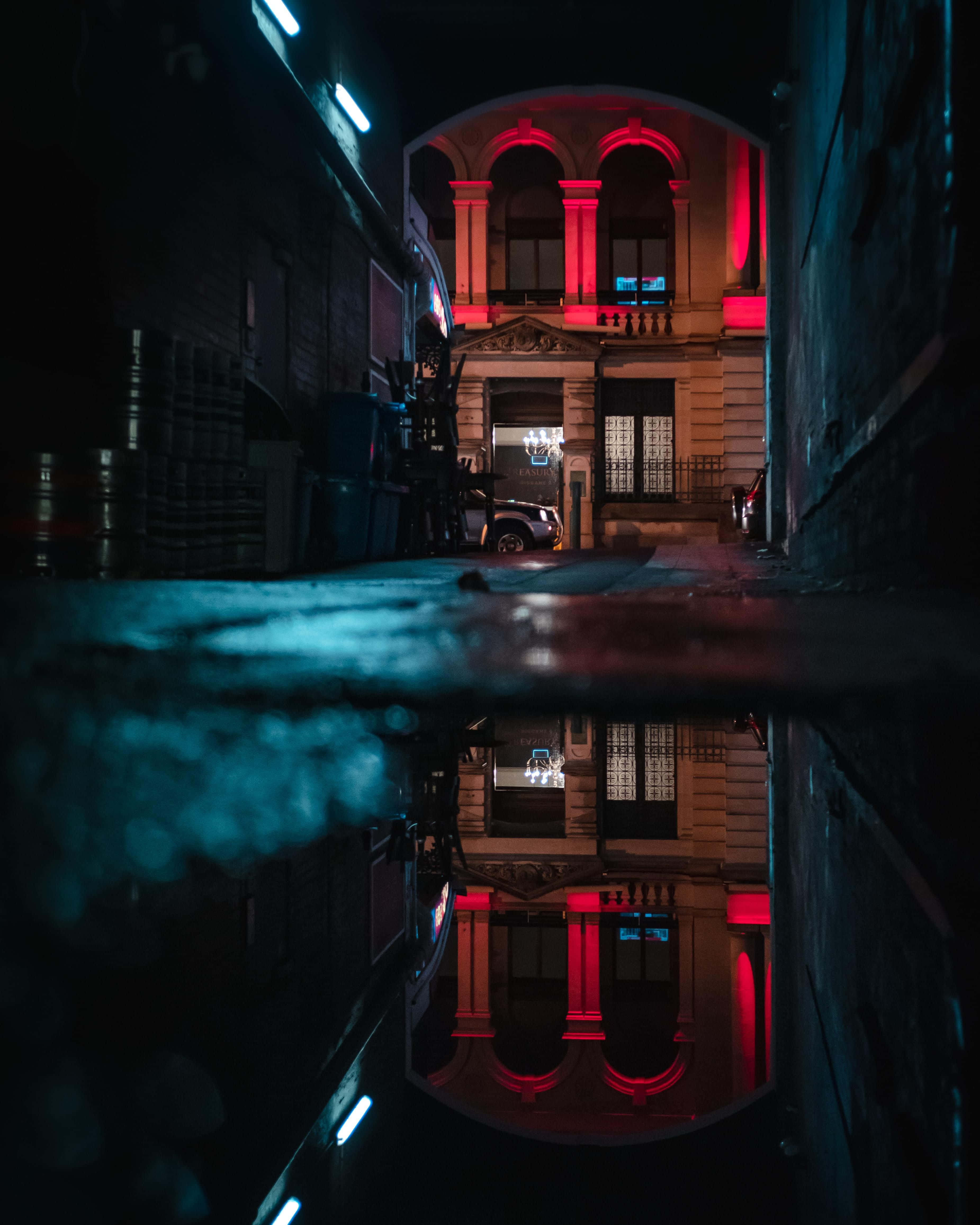 Alleyway 桌布 壁紙 — 夜晚一條狹窄的小巷，在藍色霓虹街燈下的水坑中反射出一座宏偉建築的紅色燈光外牆。