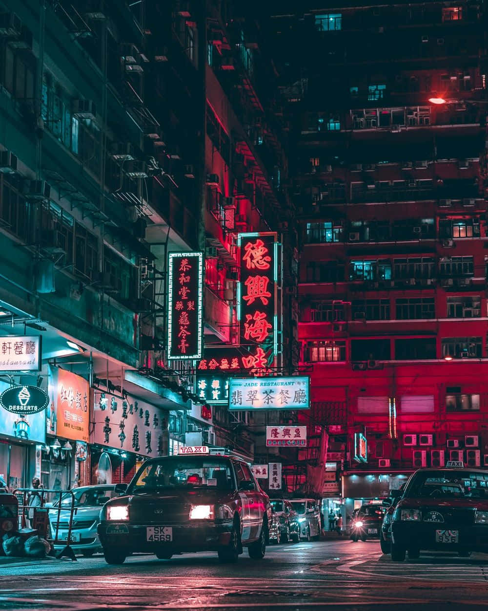 Edgy Aesthetic 桌布 壁紙 — 夜晚一條霓虹燈照亮的城市街道，紅色和藍色的招牌在潮濕的路面上反射，的士駛過高聳的建築物。
