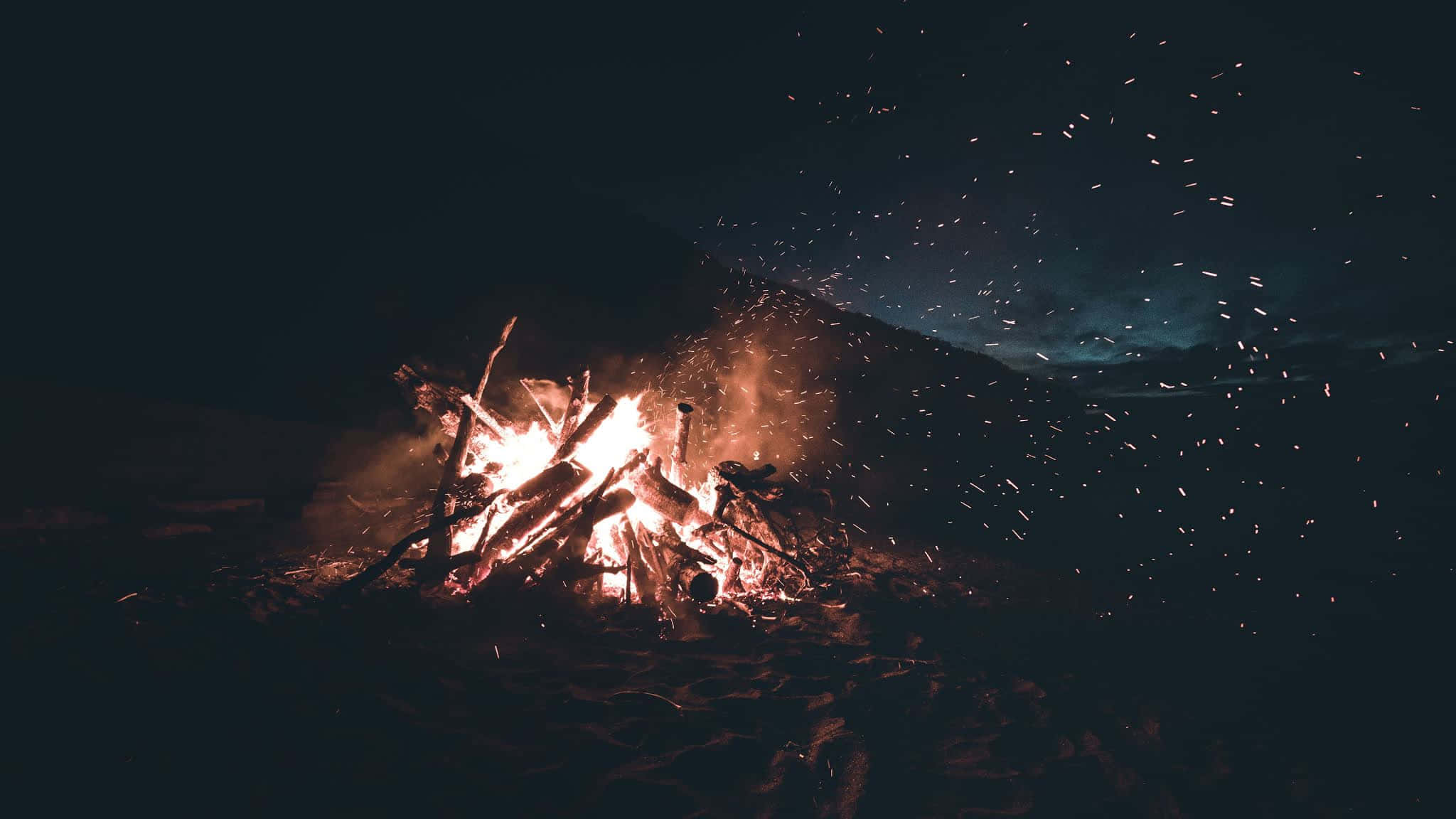 Beach Bonfire 桌布 壁紙 — 夜晚咆哮的營火將明亮的火花射向黑暗的天空，天空中有淡淡的藍色雲彩。