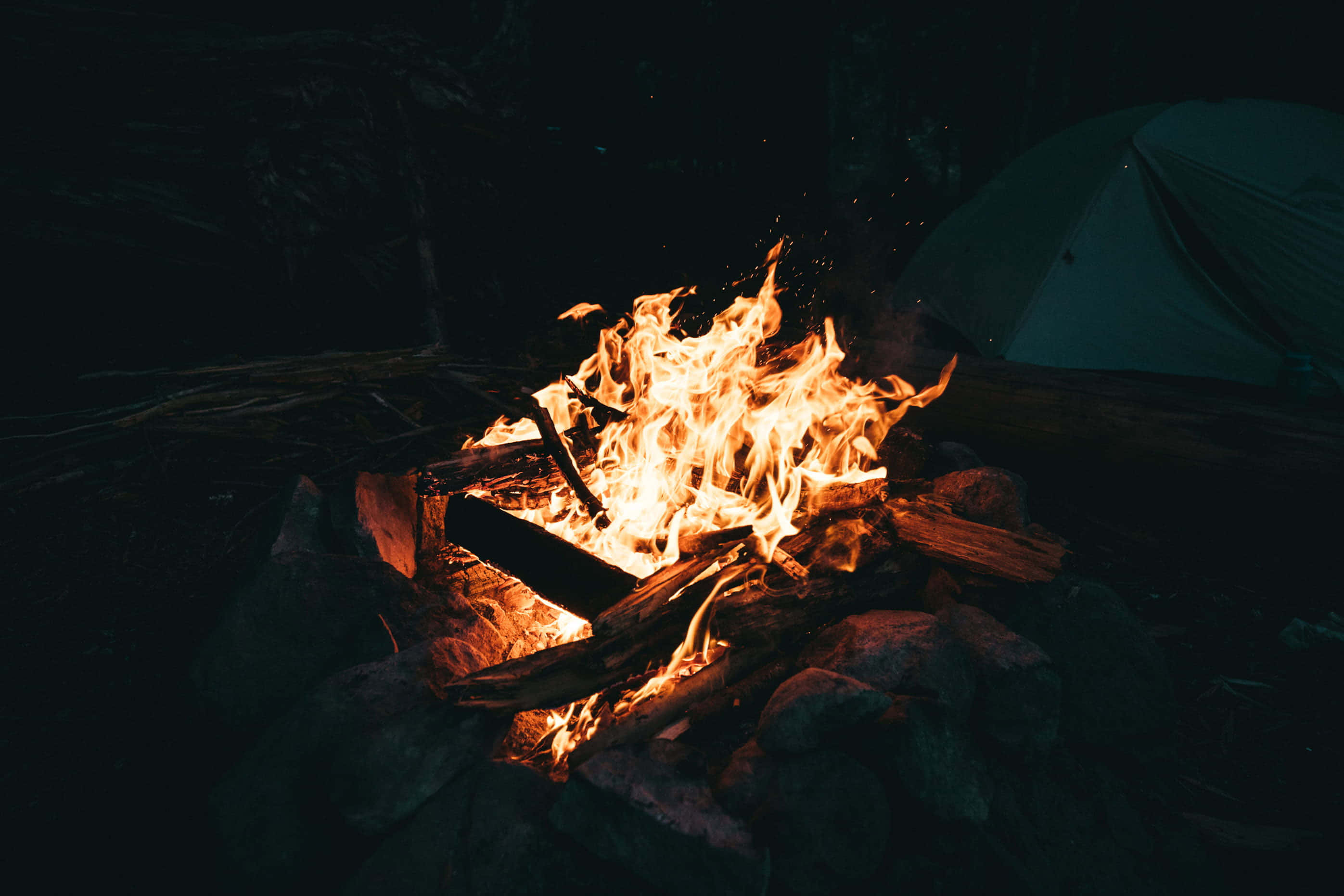 Fall Bonfire 桌布 壁紙 — 夜晚咆哮的營火照亮了黑暗荒野中的帳篷，伴隨著舞動的橙色火焰和發光的餘燼。