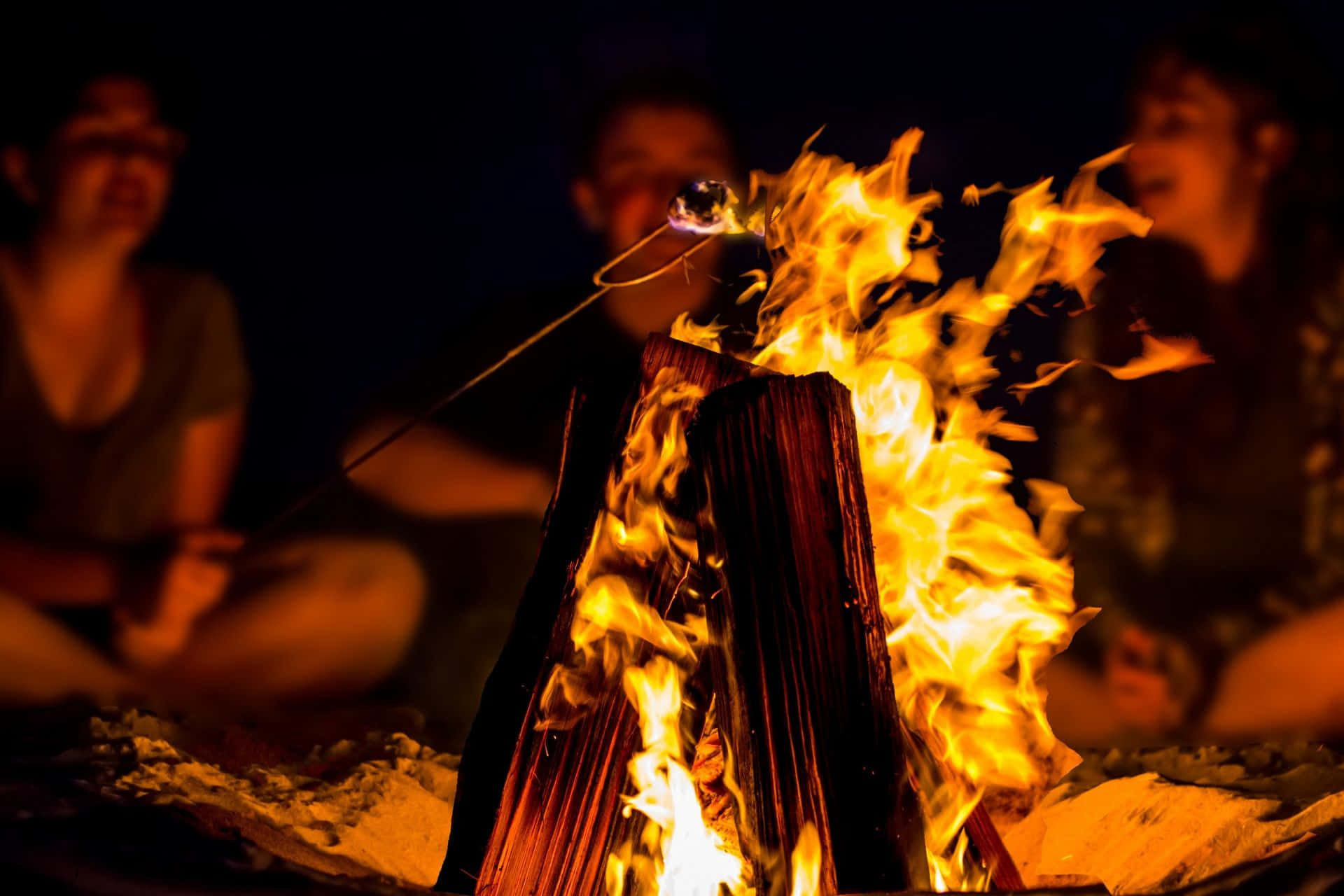 Beach Bonfire 桌布 壁紙 — 夜晚嘅營火咆哮，人們喺度烤棉花糖，發光嘅餘燼將溫暖嘅光投射喺佢哋嘅臉上。