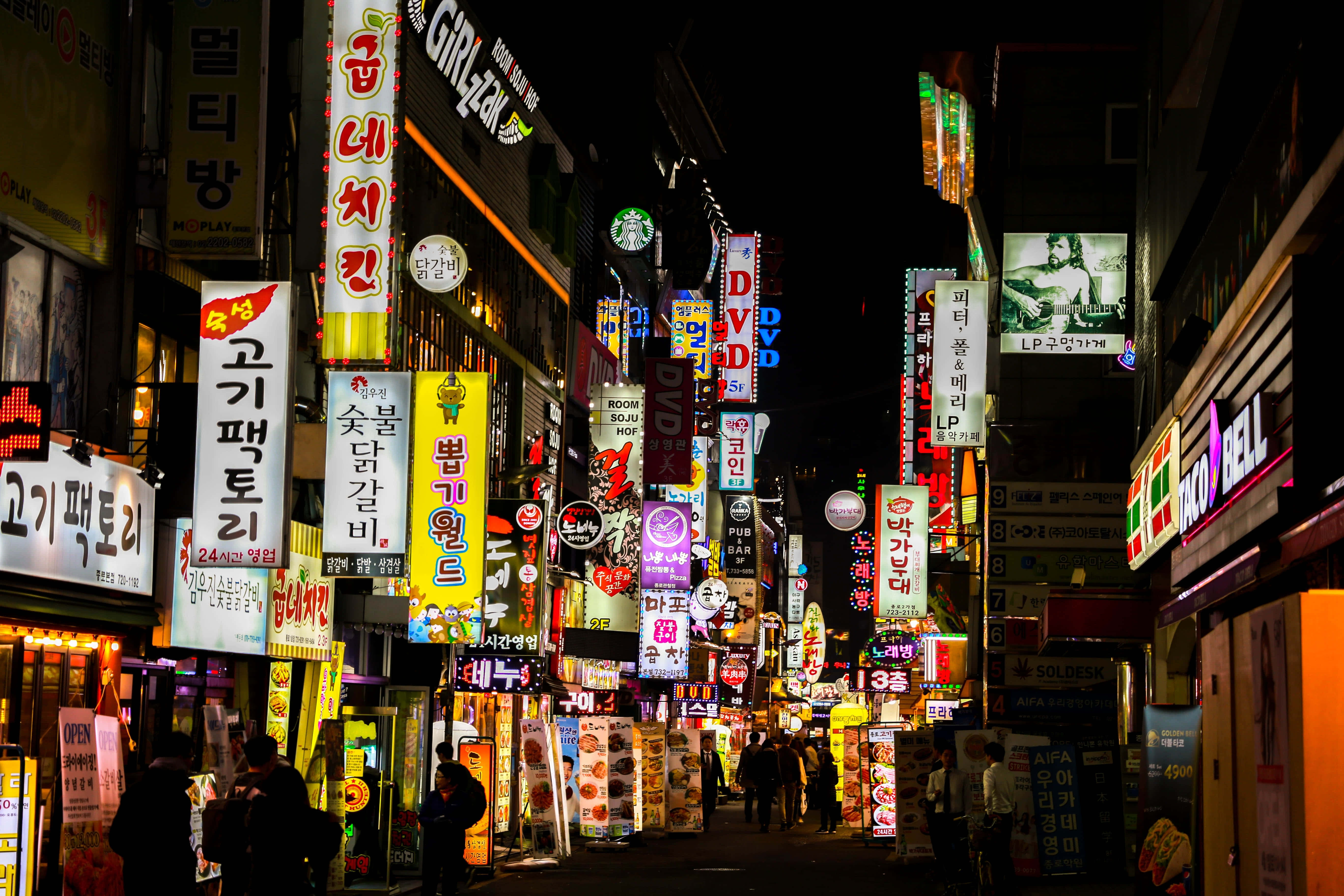 South Korea 桌布 壁紙 — 夜晚在繁華的亞洲城市中，一條閃爍霓虹燈的小巷，有發光的招牌和行人剪影。