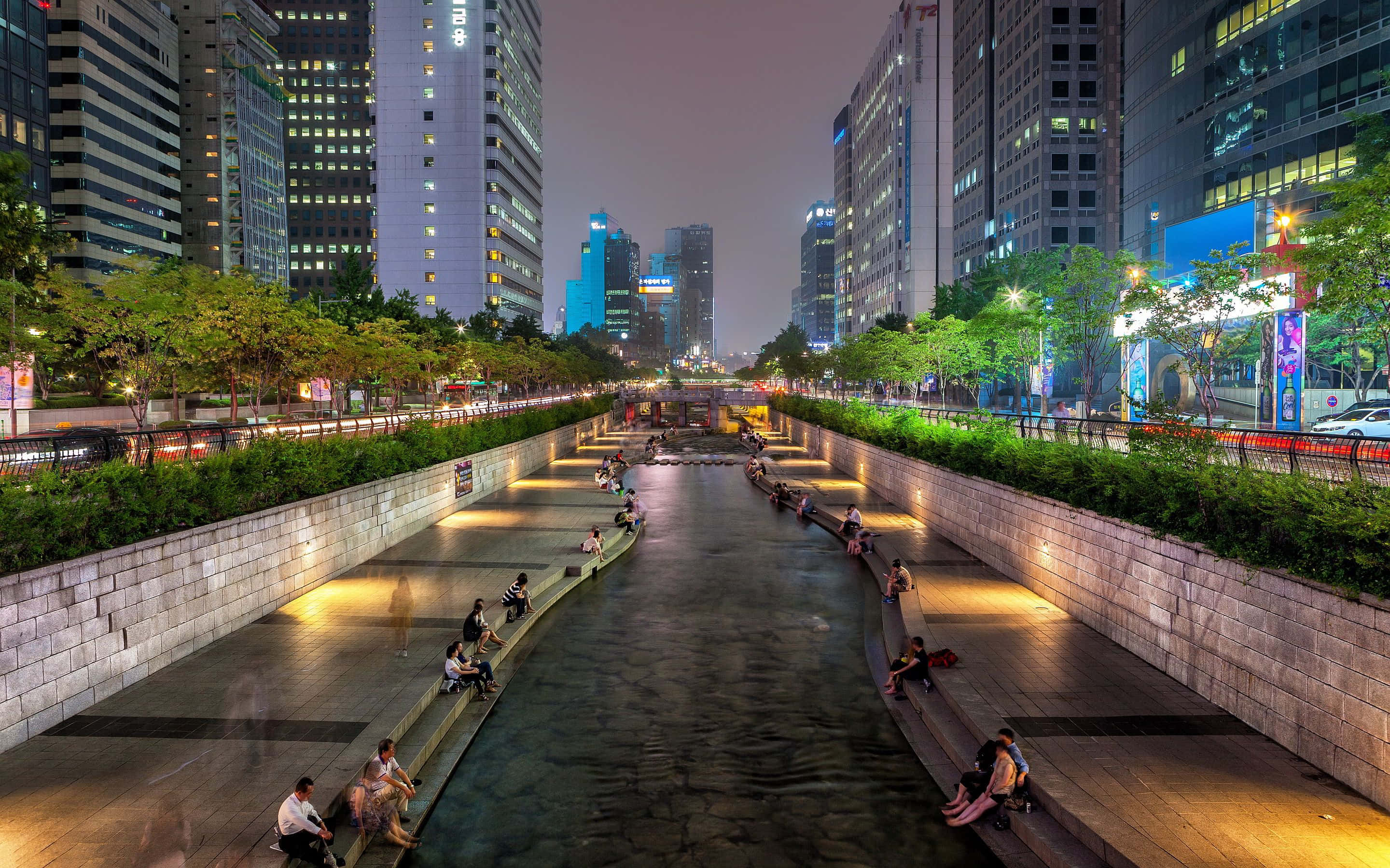 South Korea 桌布 壁紙 — 夜晚寧靜的城市運河，兩旁是閃爍的摩天大樓，人們在溫暖的路燈下於石凳上放鬆。