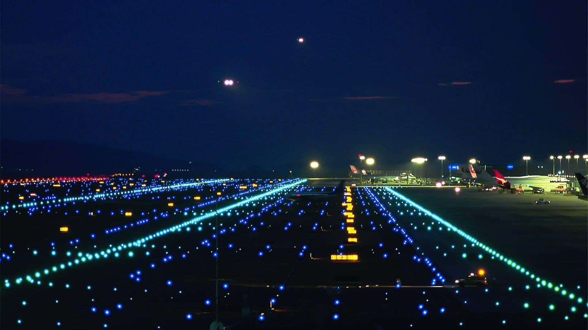 天橋 桌布 壁紙 — 夜晚從空中俯瞰機場跑道，在黑暗的天空下，藍色和黃色的燈光閃爍，遠處有飛機。