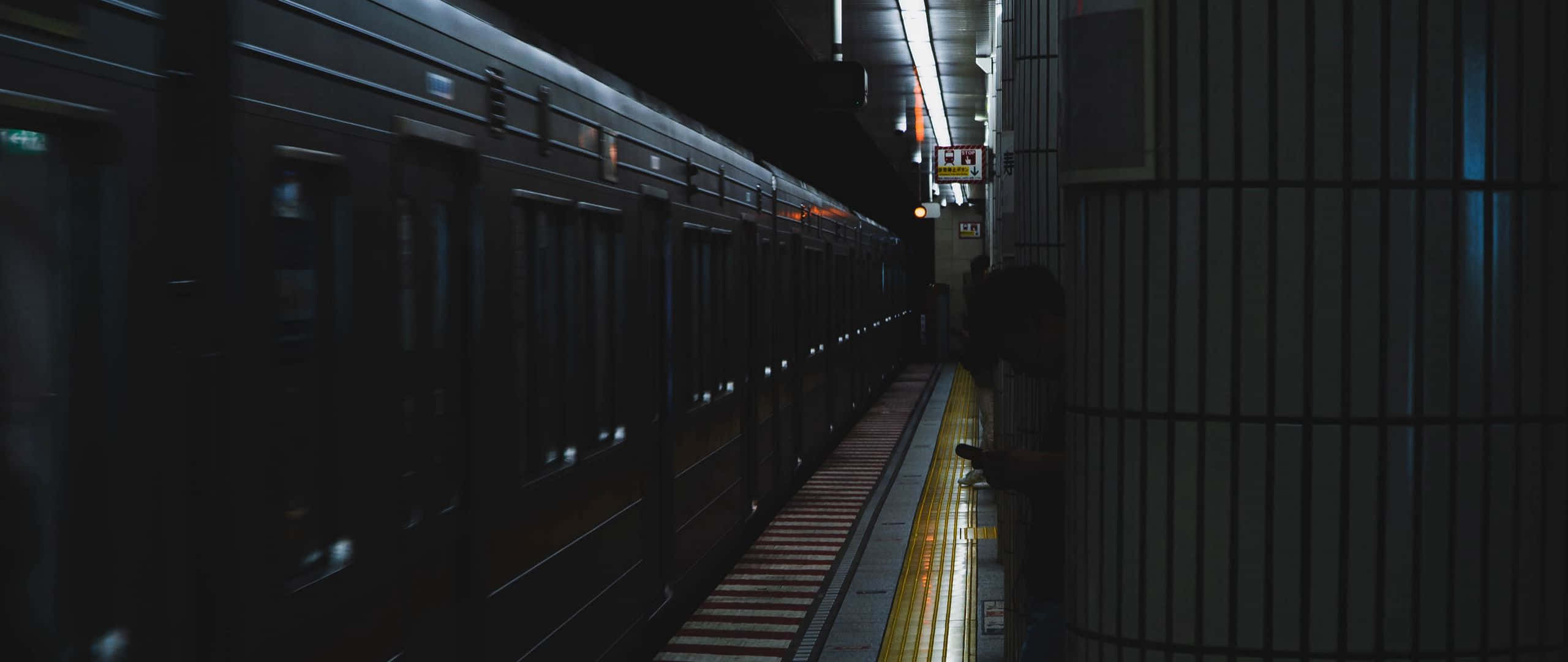 Dark Underground 桌布 壁紙 — 夜晚昏暗的地鐵月台，一列火車停在一位孤獨的乘客旁邊，該乘客站在黃色安全線附近。