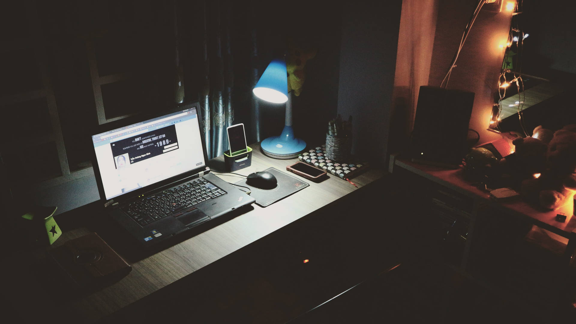 Working From Home 桌布 壁紙 — 夜晚昏暗的書桌，由藍色燈和串燈照亮，上面有手提電腦、電話和滑鼠。