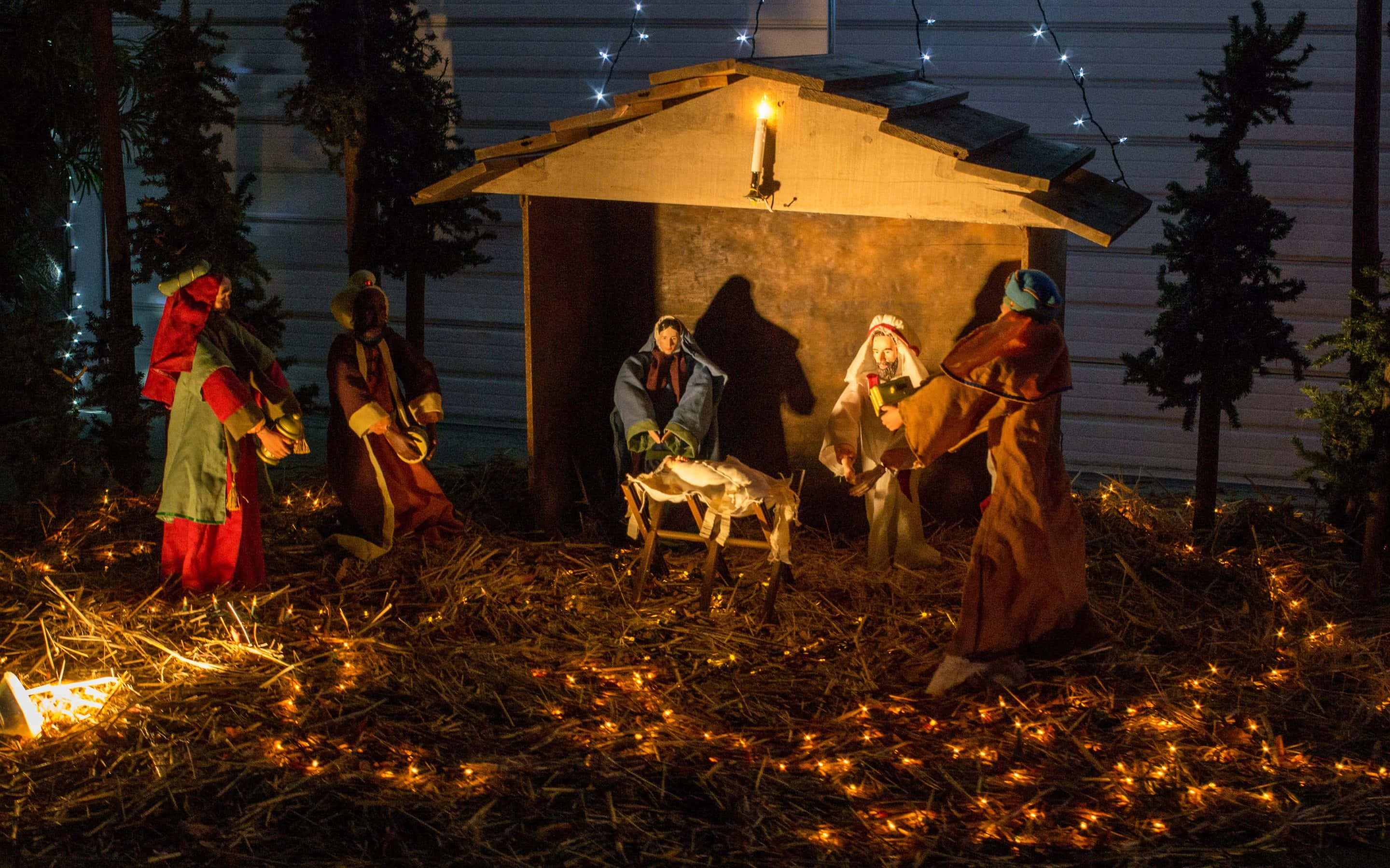 Nativity 桌布 壁紙 — 夜晚溫暖燈光下的耶穌誕生場景，馬槽周圍有雕像，被發光的仙女燈和乾草環繞。