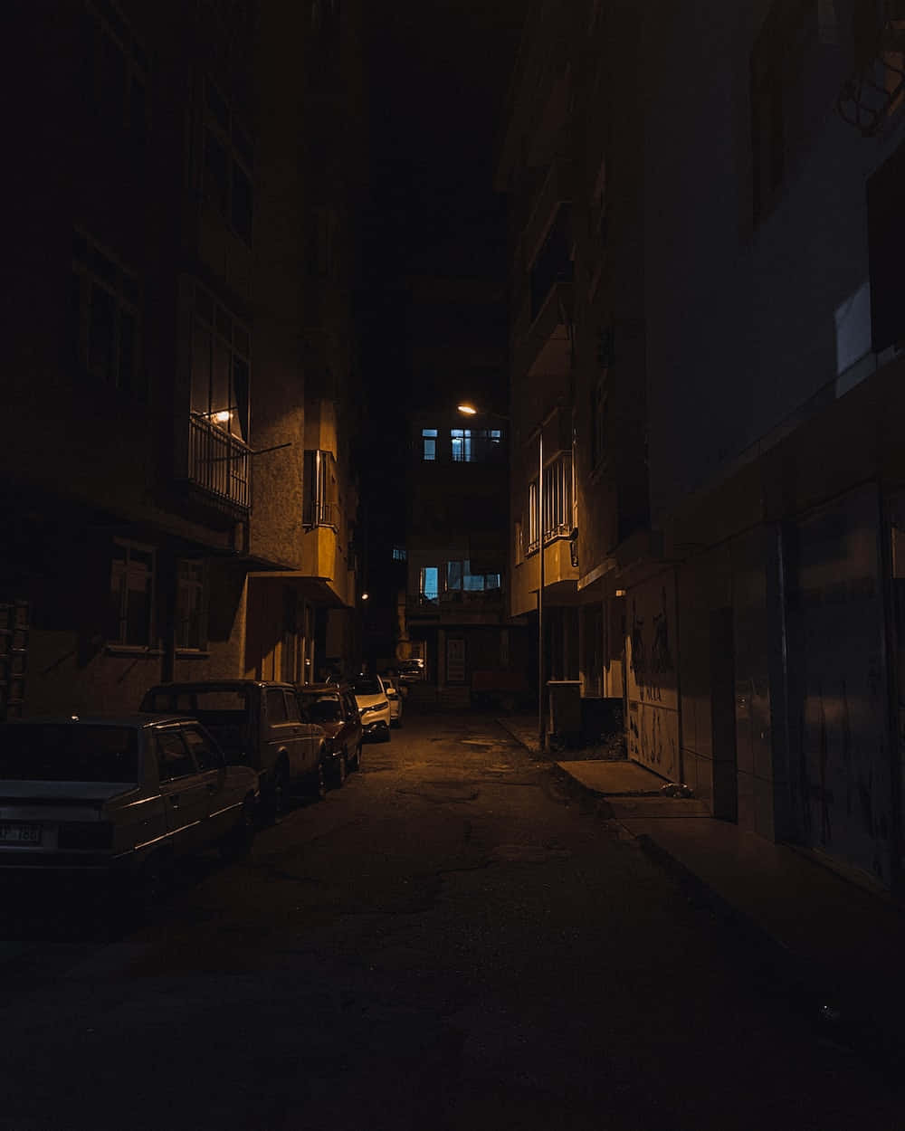 Dark Alleyway 桌布 壁紙 — 夜晚狹窄的城市小巷，溫暖的路燈和發光的窗戶在停泊的汽車上投下長長的陰影。