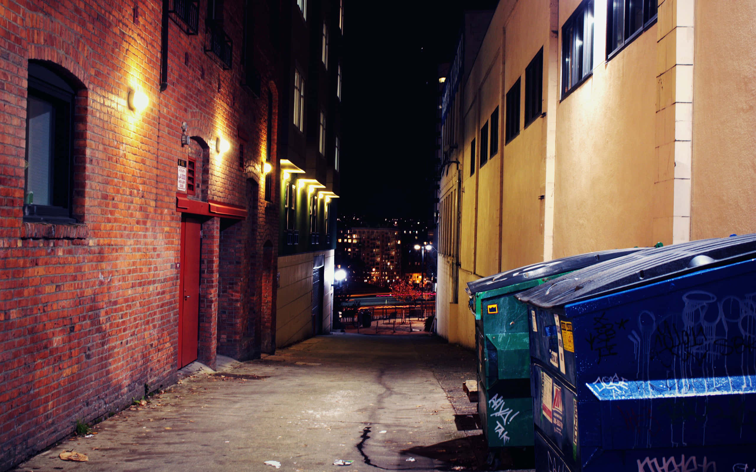 Alleyway 桌布 壁紙 — 夜晚狹窄的城市小巷，被溫暖的路燈照亮，在磚牆和塗鴉覆蓋的垃圾箱上投下長長的影子。