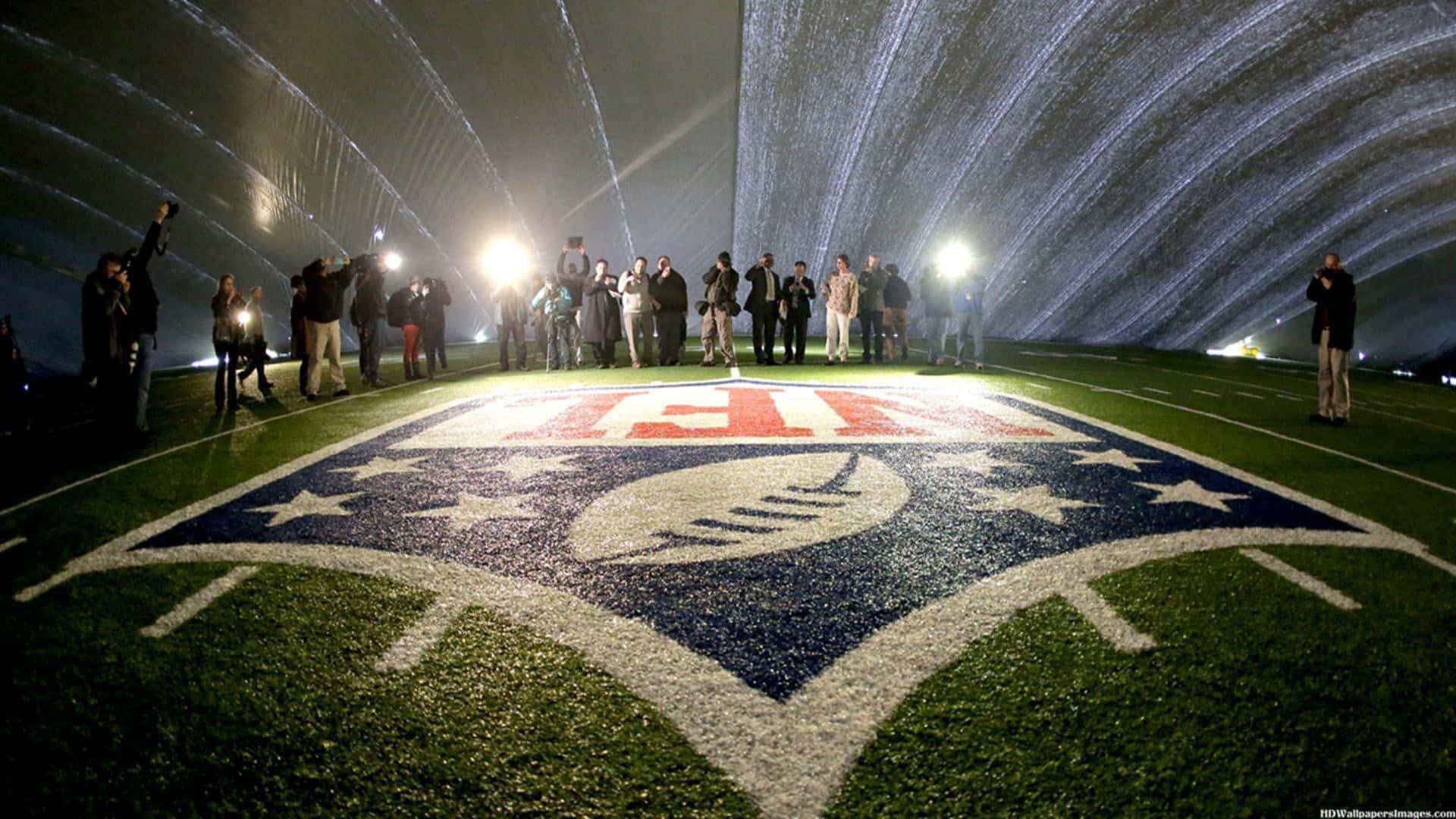 Super Bowl 桌布 壁紙 — 夜晚的足球場被明亮的燈光照亮，人們站在草地上大型球隊標誌周圍。