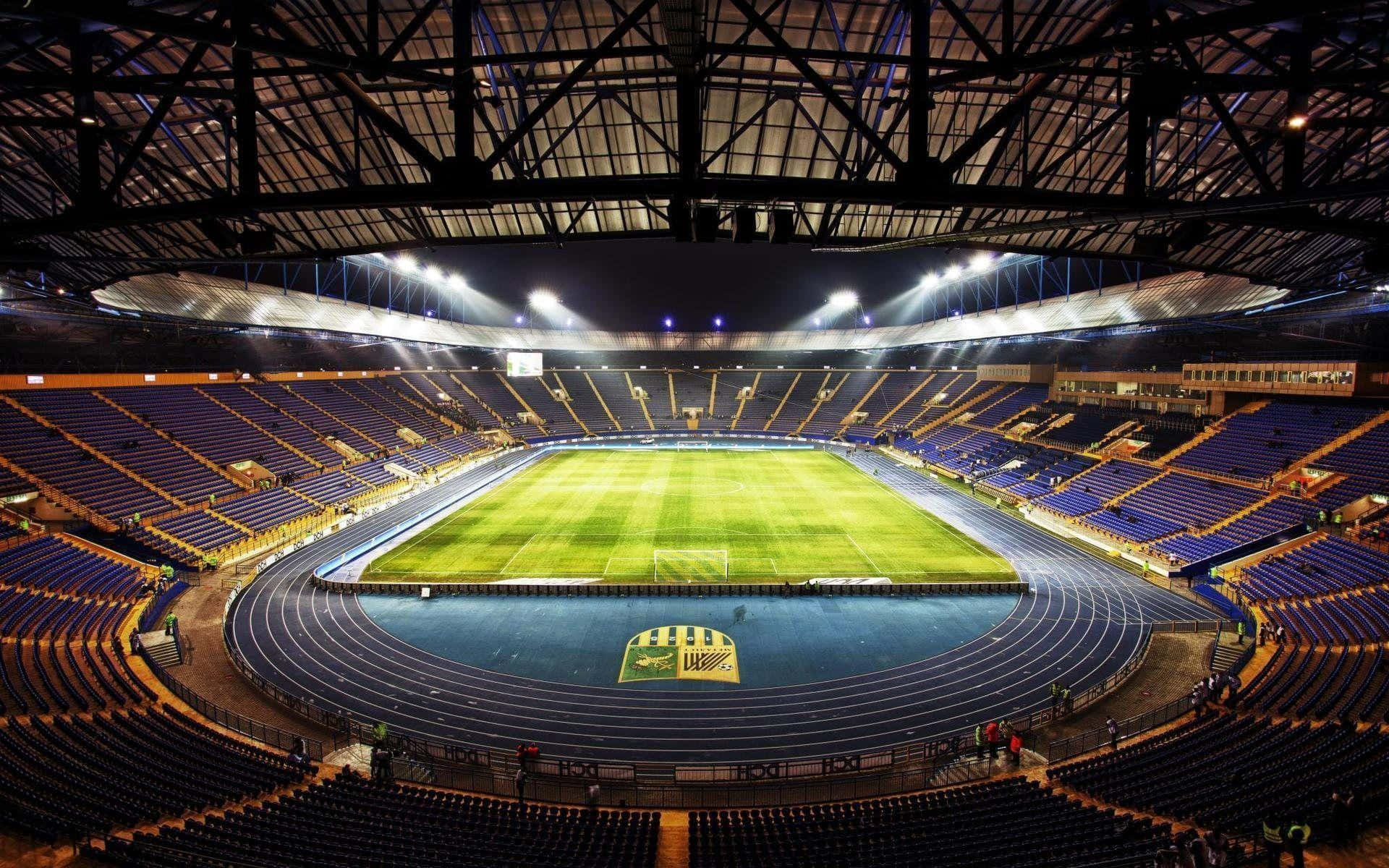 Soccer Stadium 桌布 壁紙 — 夜晚空曠大型體育館的鳥瞰圖，發光的綠色場地、藍色跑道和紫色分層座位，上方是深色屋頂。