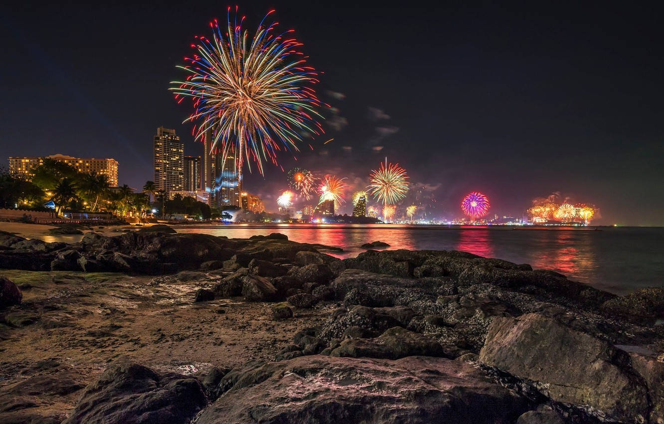 夜晚芭堤雅煙火 壁紙 — 色彩繽紛的煙花在夜間沿海城市的天際線上綻放，倒映在水面上，前景是岩石。