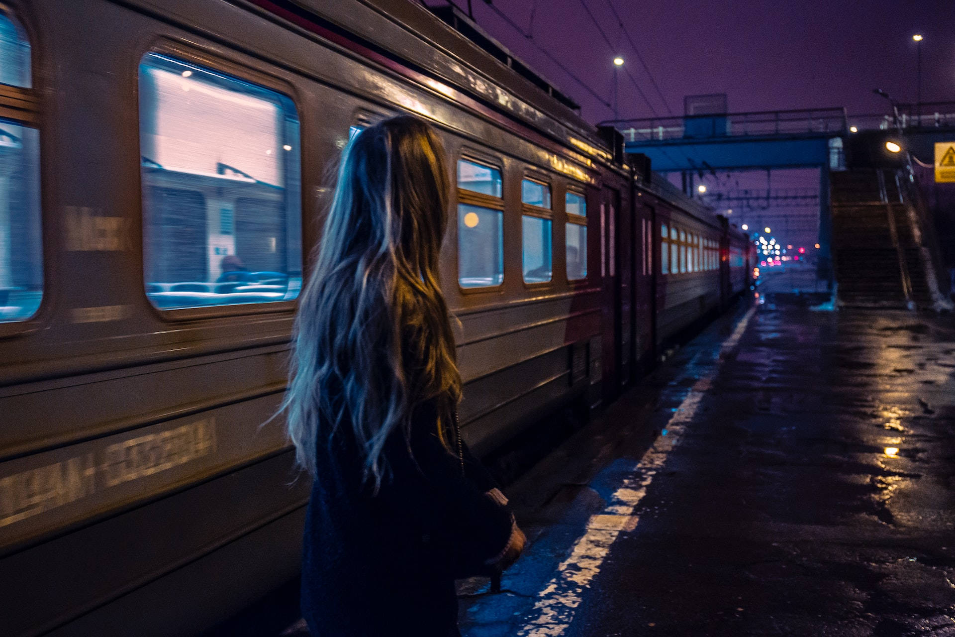 女子在濕漉漉的車站月台上看著火車，背景是紫色夜空