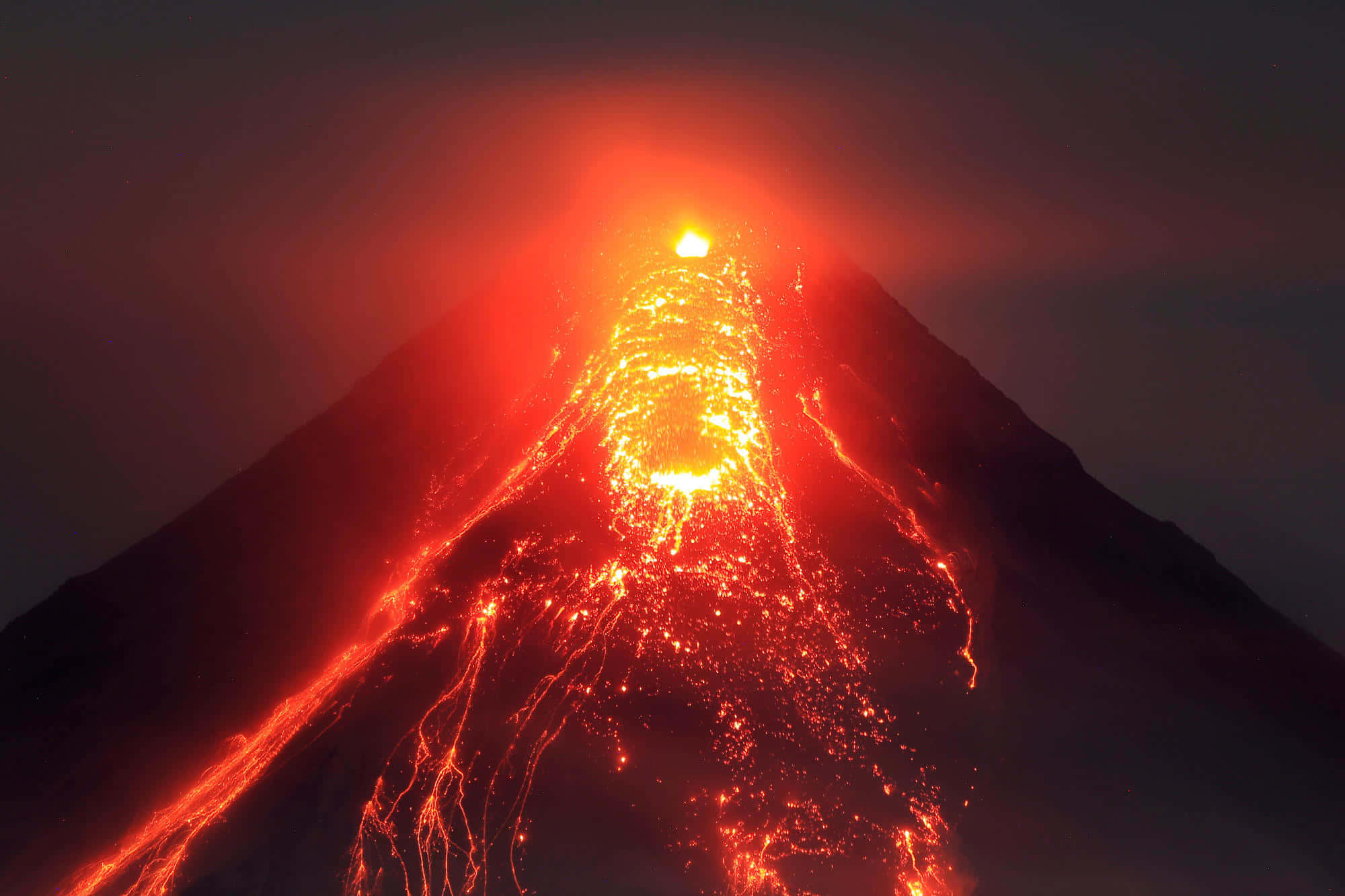 夜空下噴發的火山 壁紙 — 在漆黑的夜空下，火山在夜間爆發，熾熱的熔岩沿著山坡流下。
