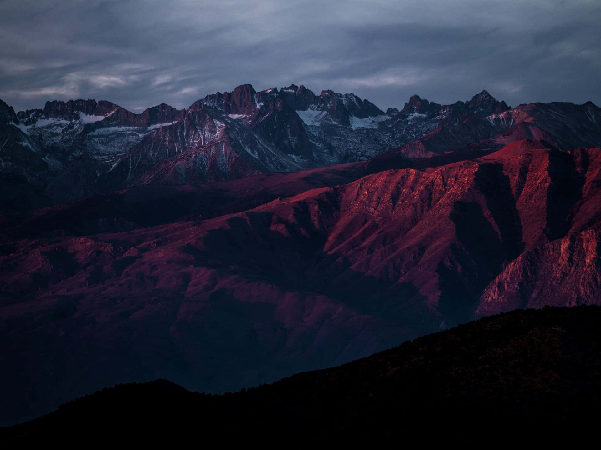 夜空下雄偉的黑暗山峰 壁紙 — 一條戲劇性的山脈沐浴在深紅色的暮色中，白雪覆蓋的山峰在陰沉的灰色天空下。