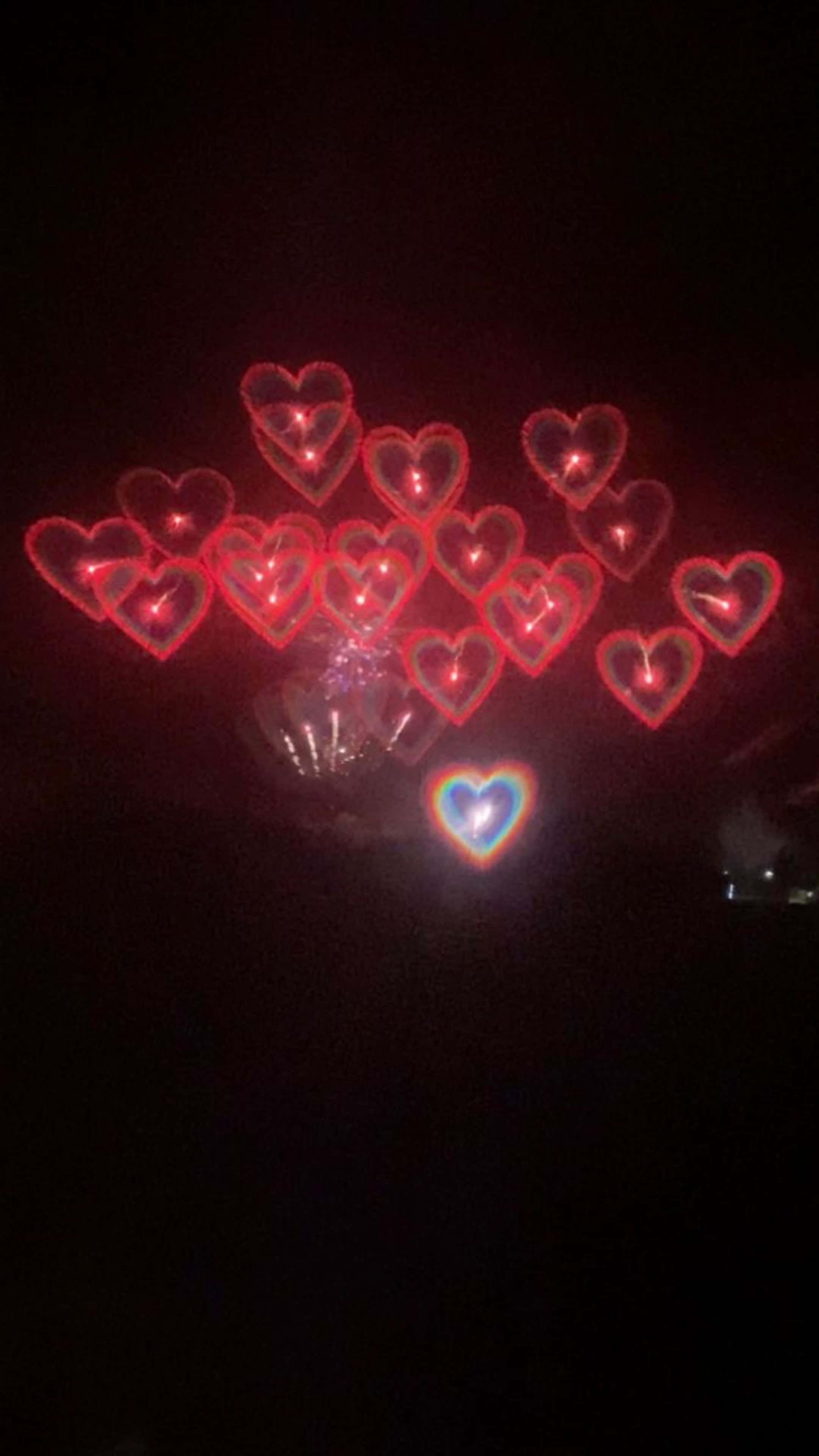 夜空中心形煙火 壁紙 — 一簇發光的紅色心形燈光在漆黑的夜空中飄浮，下方有一顆彩虹心形。