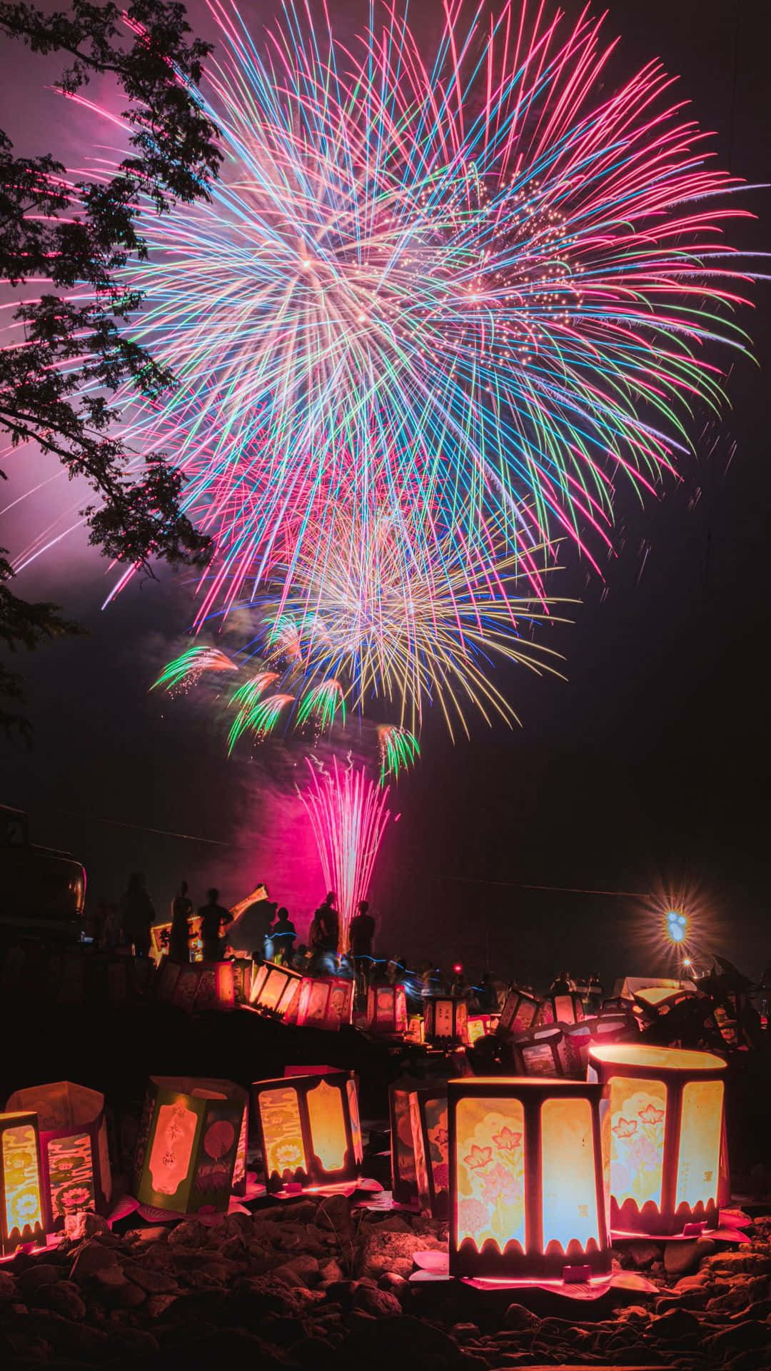 夜空的璀璨煙火 壁紙 — 色彩繽紛的煙火在粉紅、藍和綠色中爆炸，在發光的燈籠上方，剪影觀眾在夜晚從岩石山坡觀看。