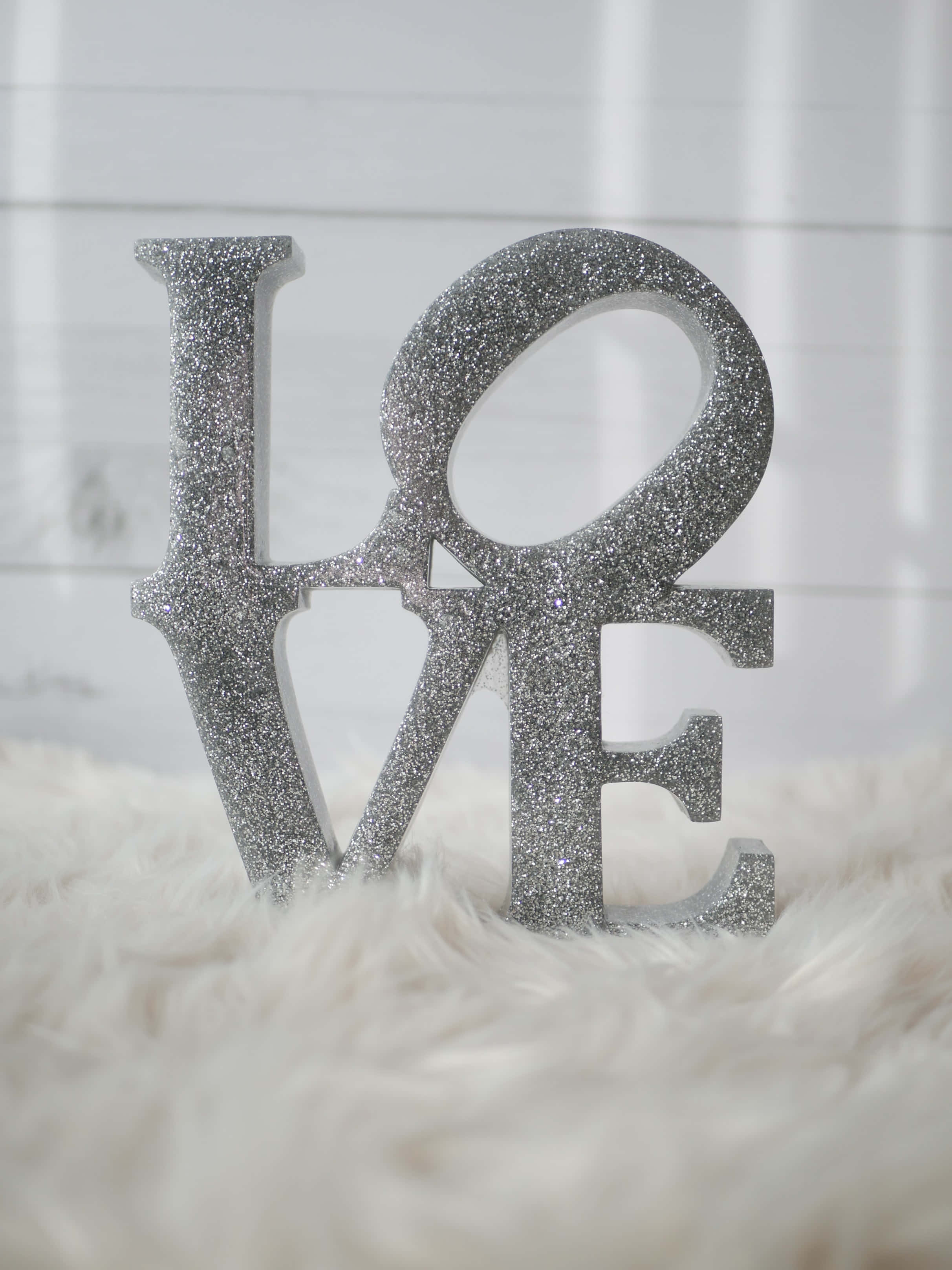 夜空背景上的閃耀銀色星光 壁紙 — 一個閃亮的銀色「LOVE」字雕塑優雅地立在白色毛絨地毯上，背景是明亮的白色護牆板牆壁。