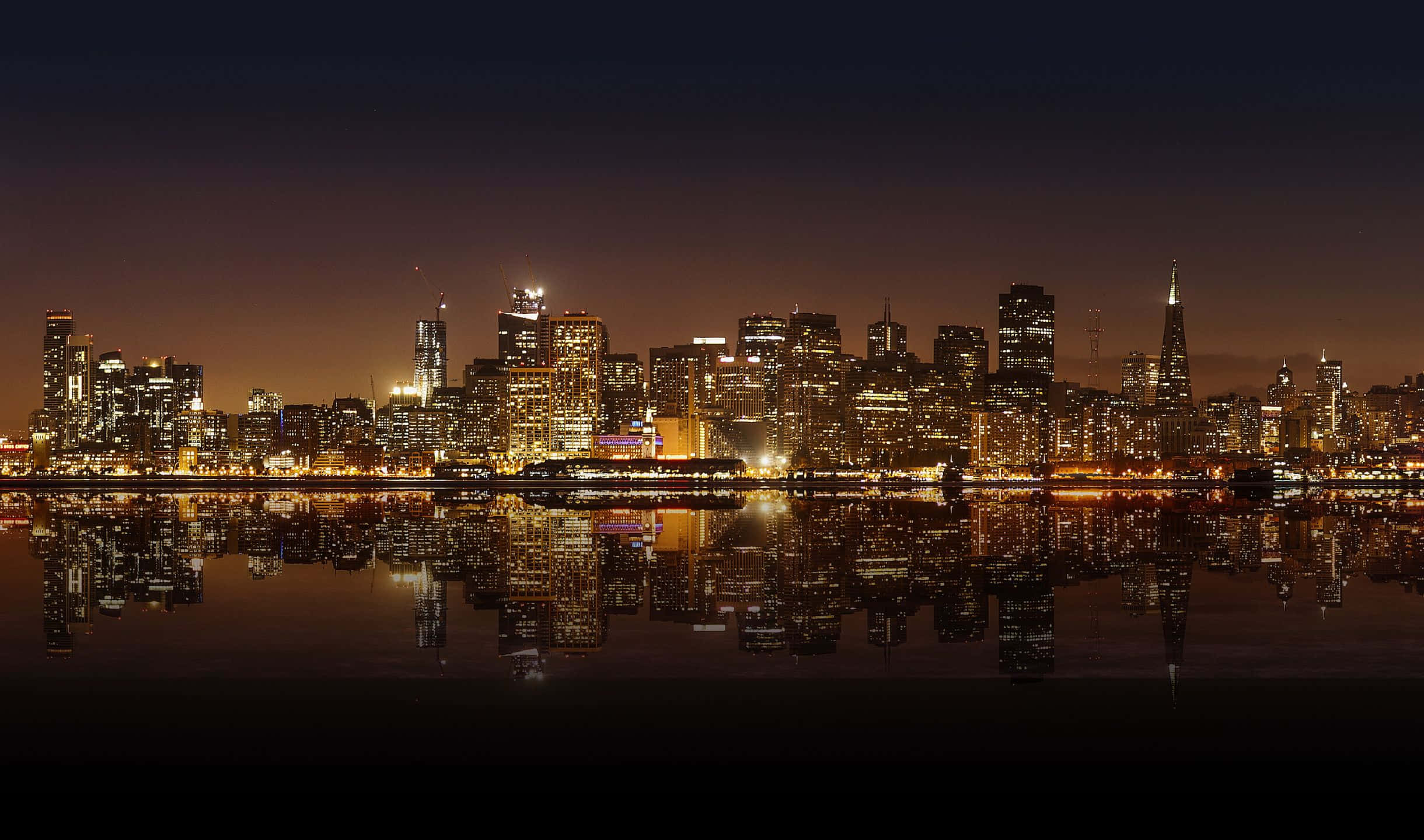 夜色中照亮的灣區大橋與舊金山天際線 壁紙 — 城市天際線的 panoramic 夜景，發光的摩天大樓完美倒映在平靜的水面上。