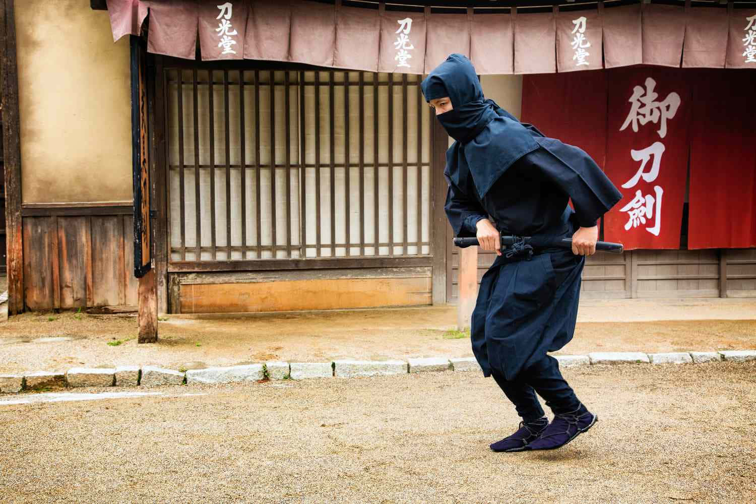 身穿深藍色裝備的忍者，在傳統日本建築前手持劍。