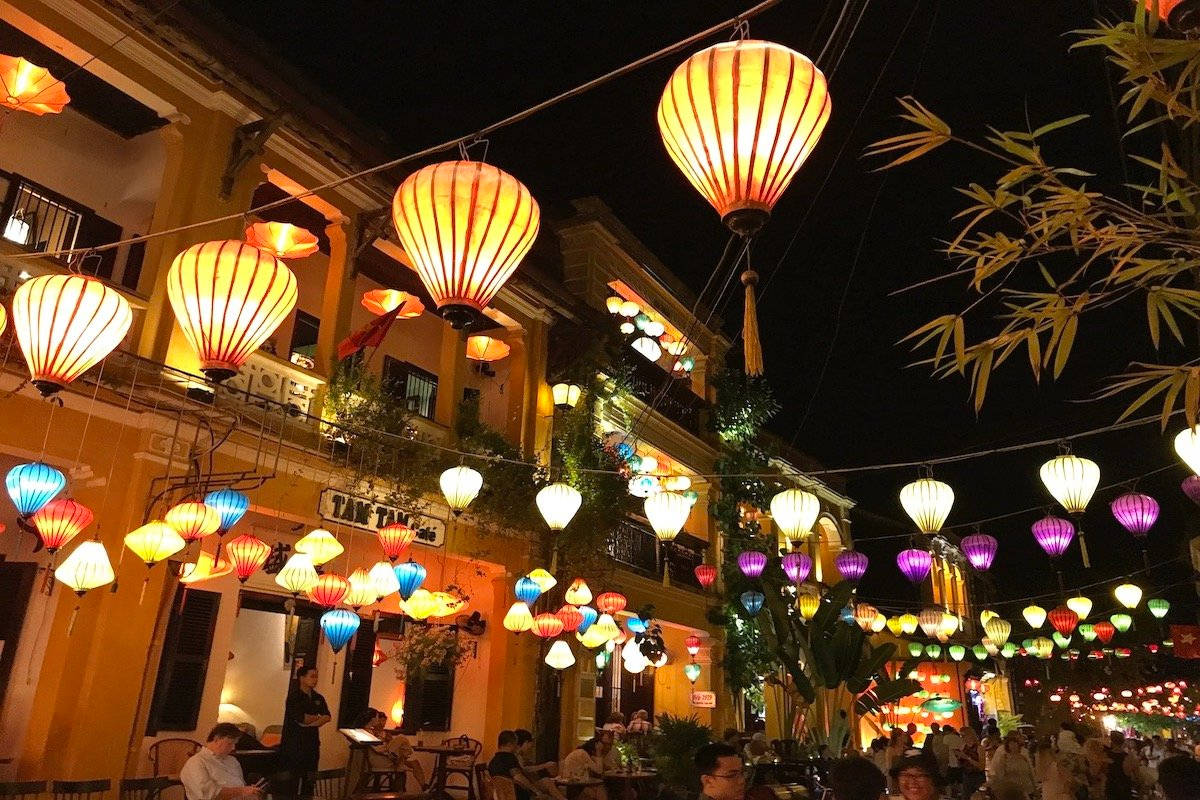夜色中的河內彩燈 壁紙 — 鮮豔的紙燈籠在黑暗的夜空下溫暖地發光，照亮了一條兩旁種滿黃色建築物和竹樹的繁華街道。