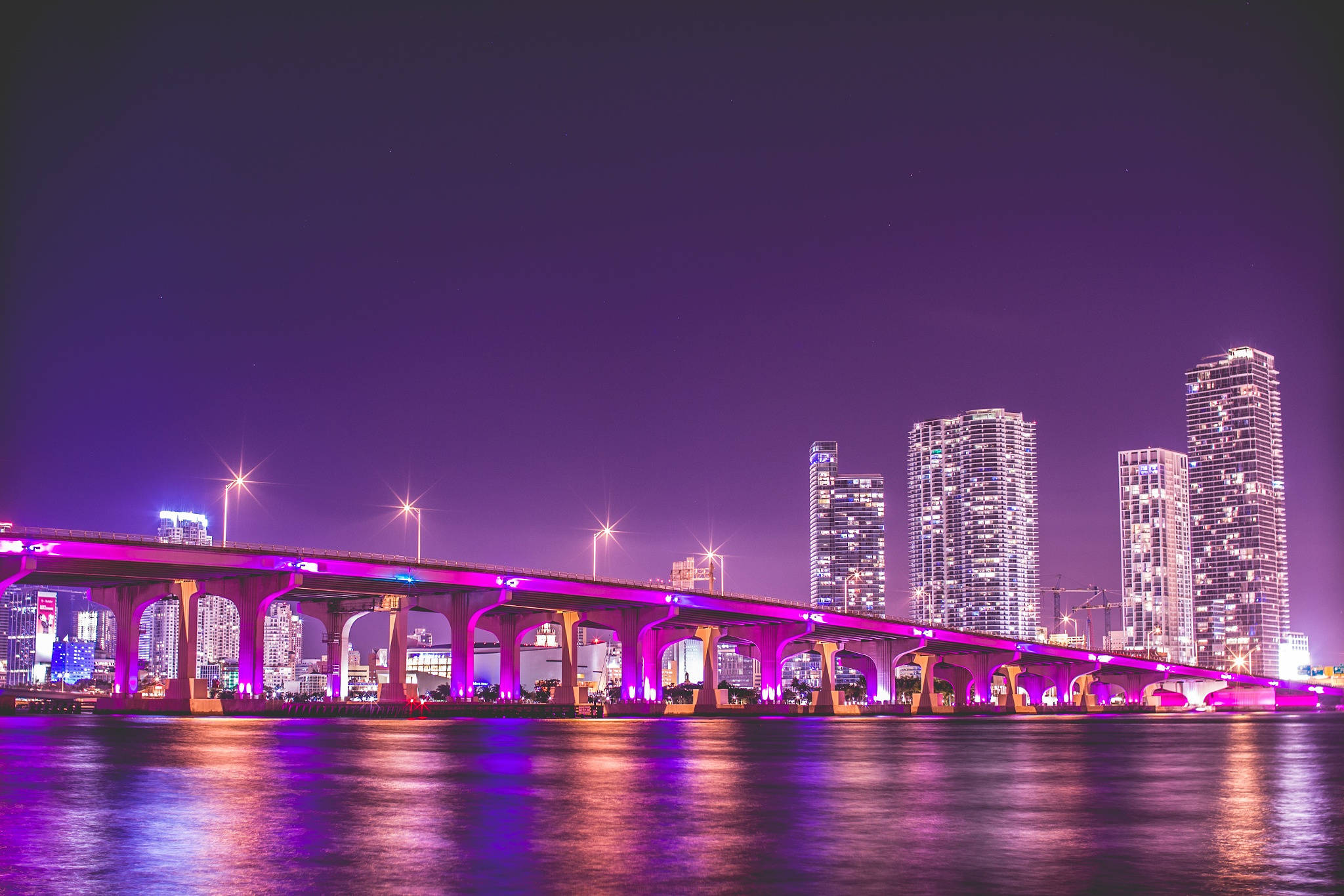夜色中色彩繽紛的邁阿密橋 壁紙 — 一座充滿活力的紫色燈光橋樑橫跨平靜的河流，夜晚中發光的摩天大樓倒映在水面上。