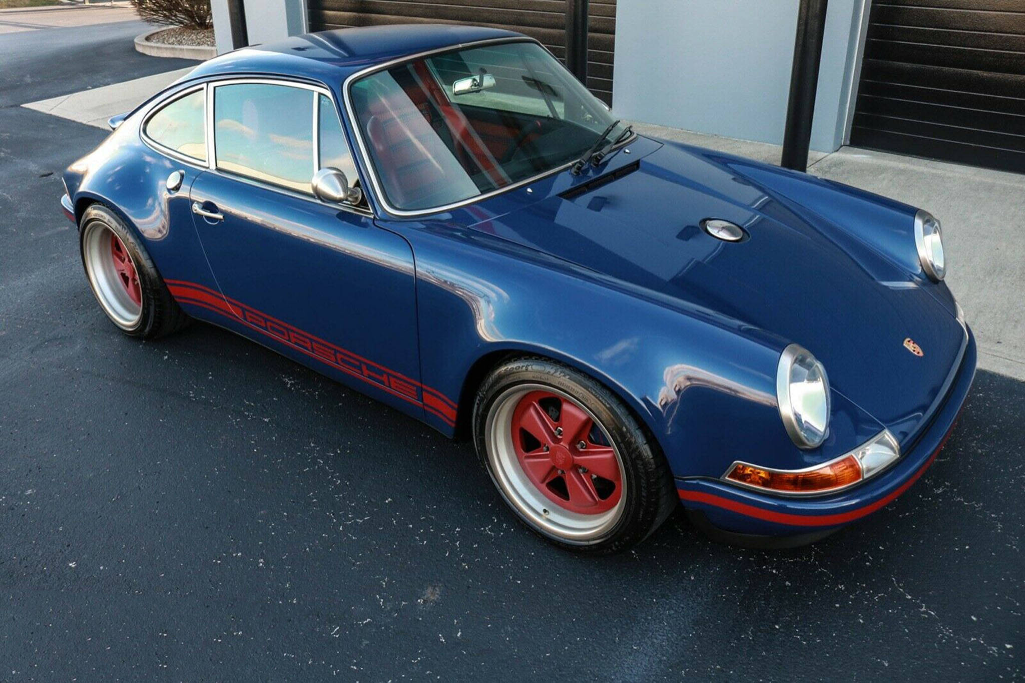 夜藍Singer Porsche 壁紙 — 一輛帶有紅色點綴和輪轂的光澤藍色保時捷911停在建築物附近的瀝青路面上。