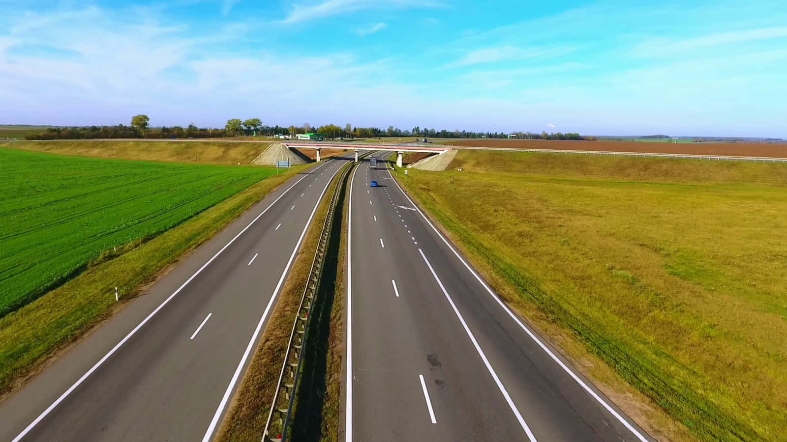 夜行公路的孤寂之美 壁紙 — 從高空俯瞰，一條寬闊的高速公路在晴朗的藍天下穿過充滿活力的綠色和金色田野。