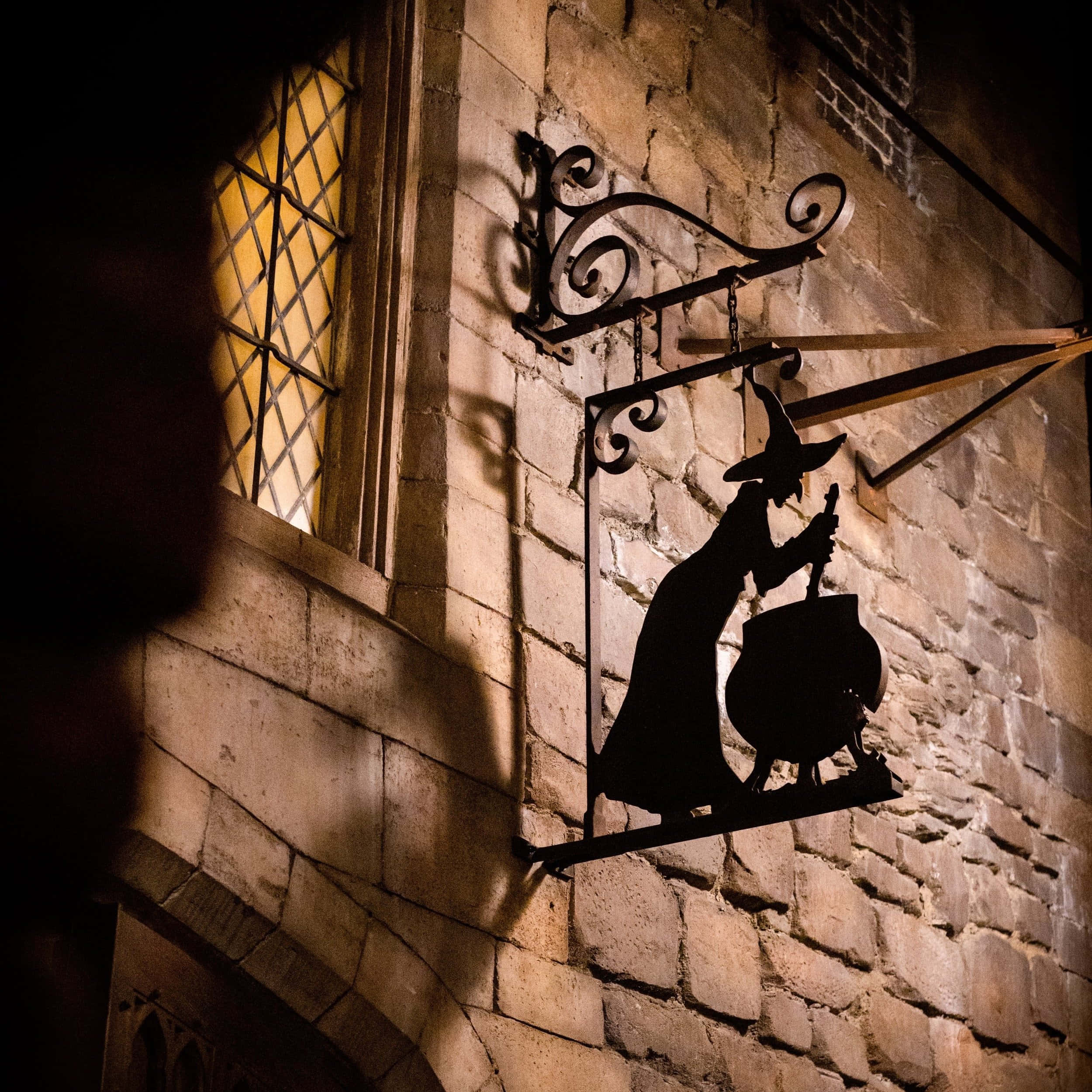 The Leaky Cauldron 桌布 壁紙 — 夜間，一個女巫剪影標誌掛在石牆上，被菱形窗戶透出的溫暖燈光照亮。