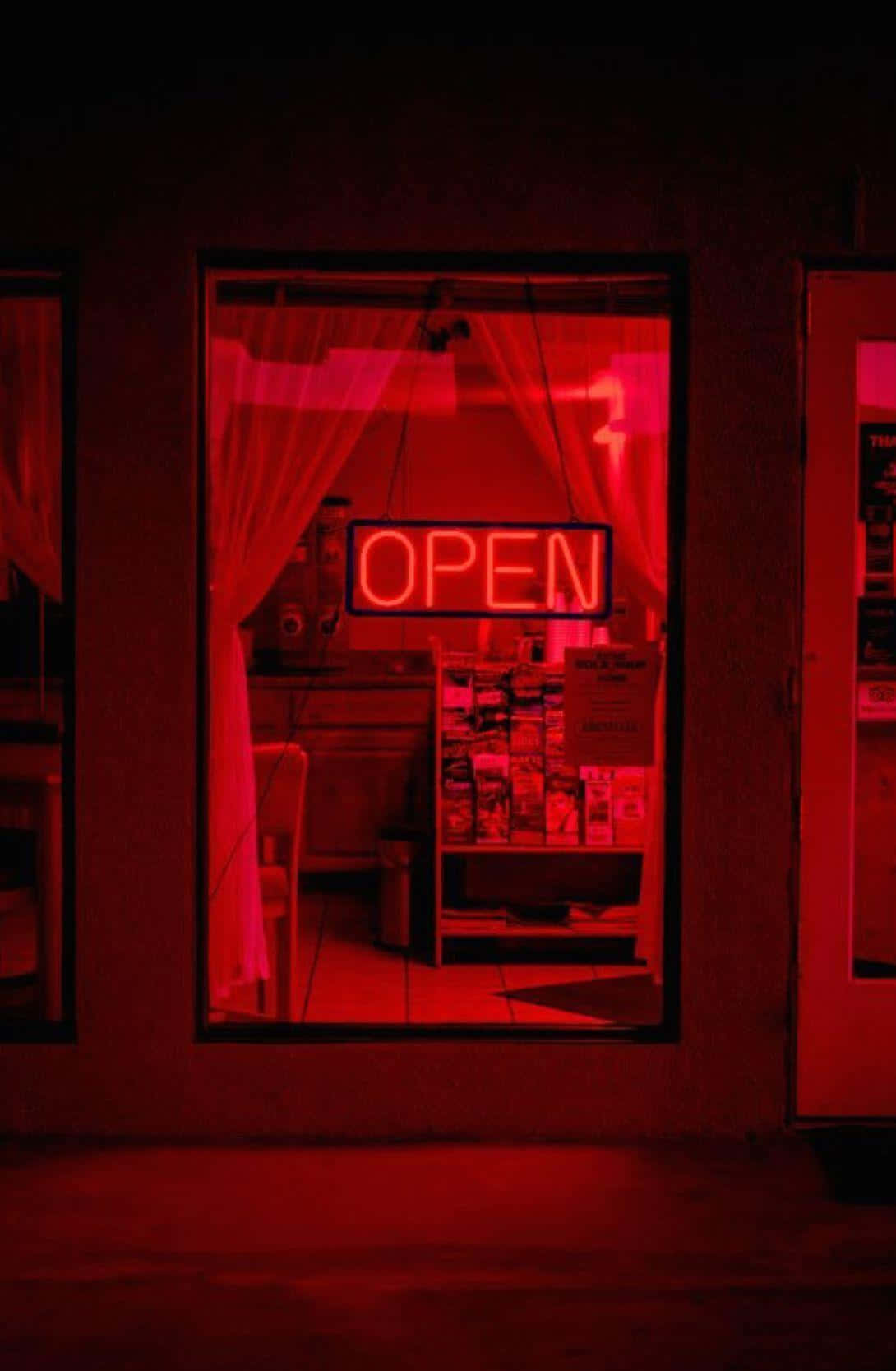Pastel Red Aesthetic 桌布 壁紙 — 夜間，一個發光的紅色霓虹燈「OPEN」標誌透過玻璃窗照亮了光線昏暗的店舖內部。
