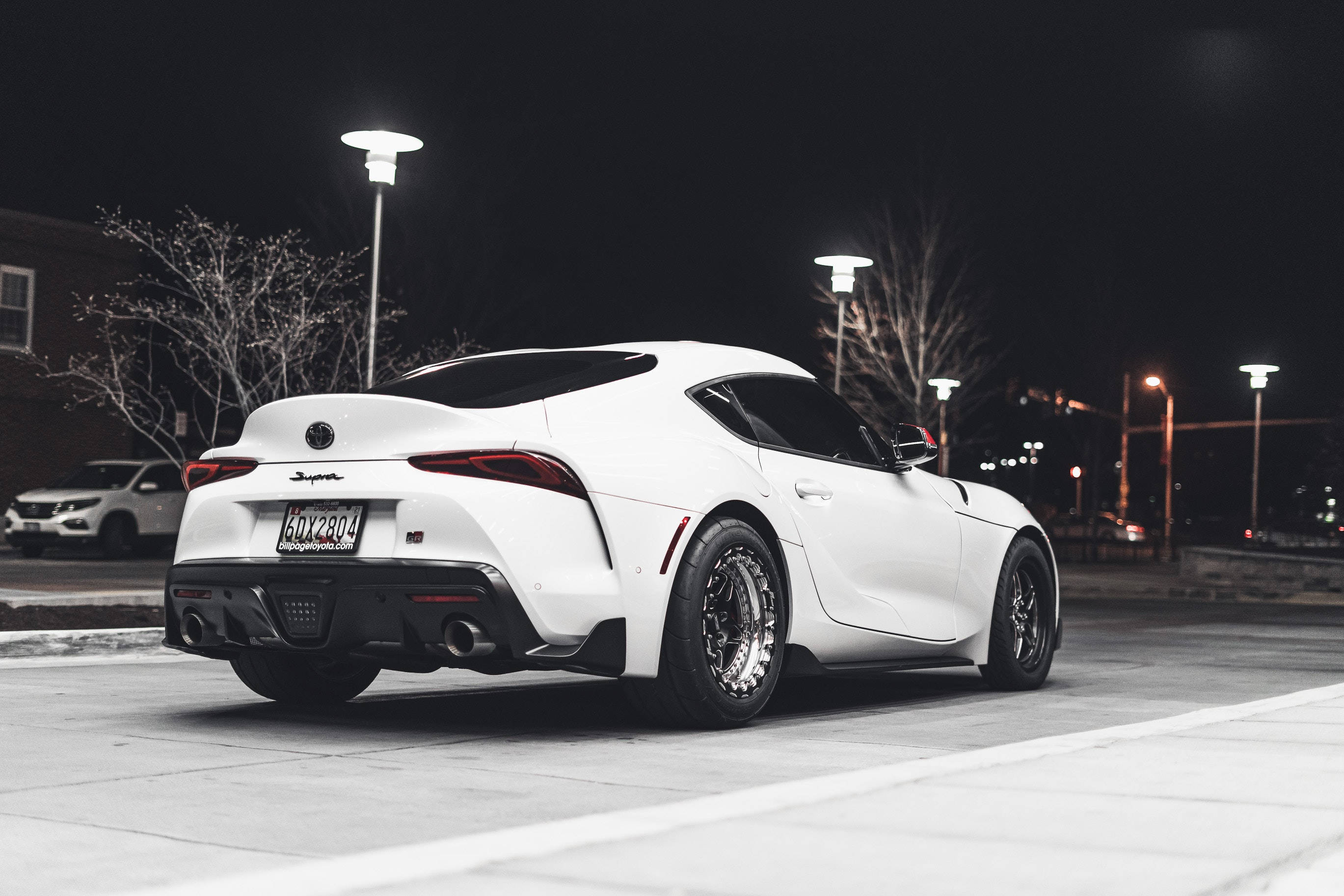夜間4K白色Toyota Supra 壁紙 — 一輛時尚的白色跑車在夜晚停泊在街燈下，背景是光禿的樹木。