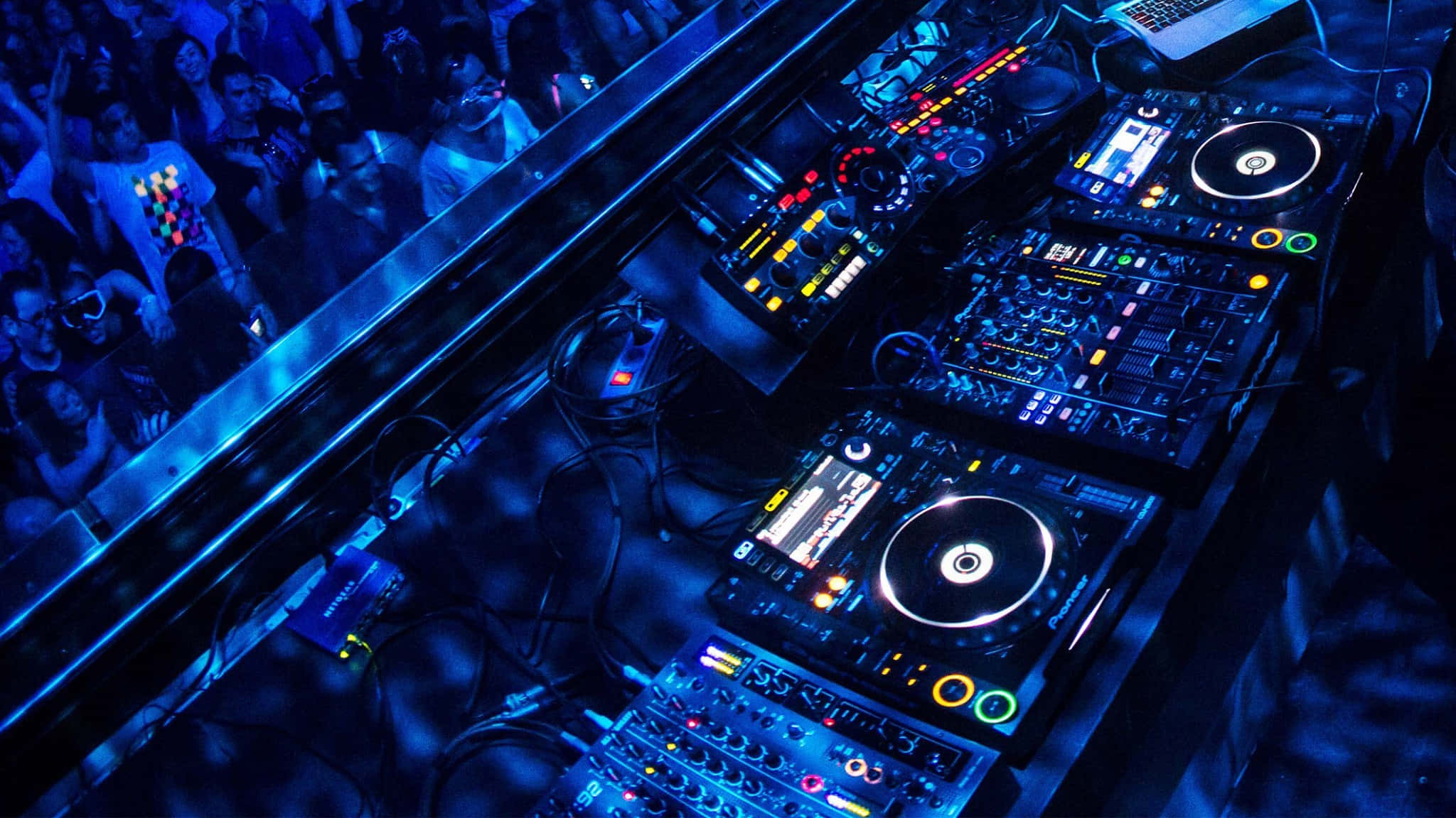 夜間DJ音樂設置圖 壁紙 — Overhead view of glowing DJ equipment bathed in blue light with a crowd watching below.