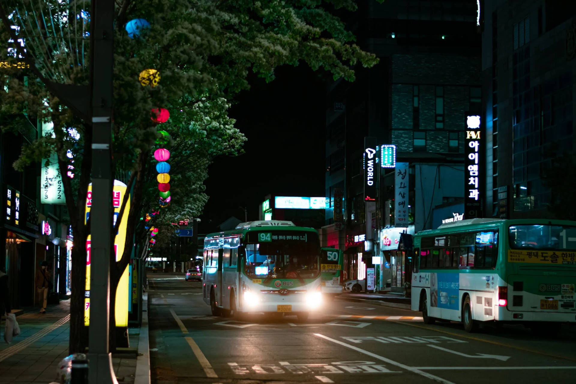 夜晚城市街道，有巴士和發光的霓虹燈招牌，樹下掛著燈籠。