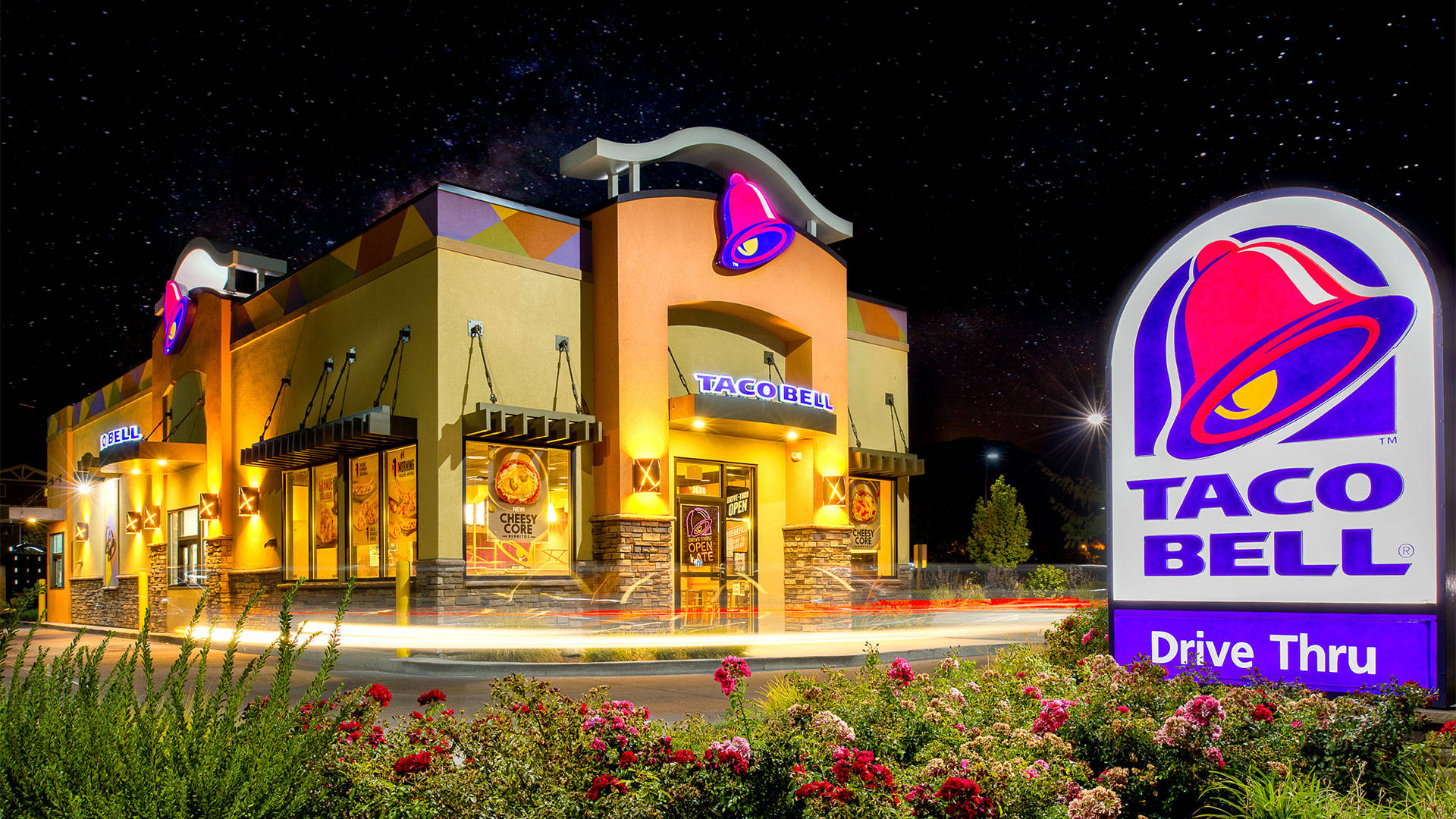 夜間塔可鐘得來速 壁紙 — 夜晚星空下，一間燈火通明的Taco Bell餐廳，發光的招牌和動態模糊的車輛駛過。