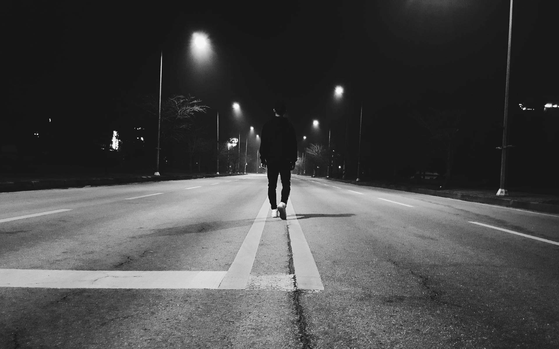 夜間孤身立於空曠道路的男子 壁紙 — 一個孤獨的身影在夜晚沿著一條荒涼的街道行走，被路燈照亮，投下長長的影子。