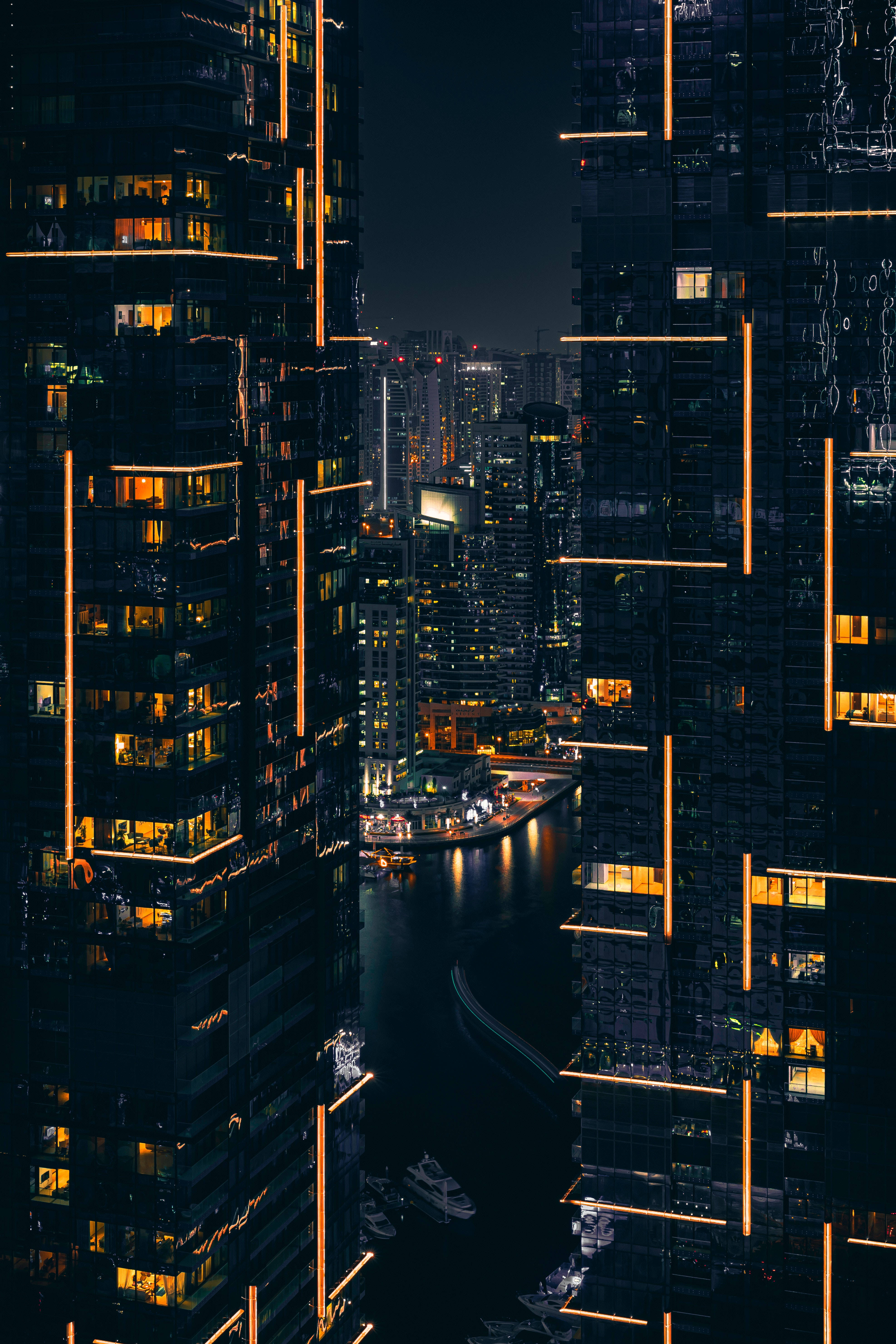 夜間建築燈光 壁紙 — 夜晚的城市景觀，由高聳的摩天大樓框住，琥珀色的燈光在下方黑暗的水道上反射。